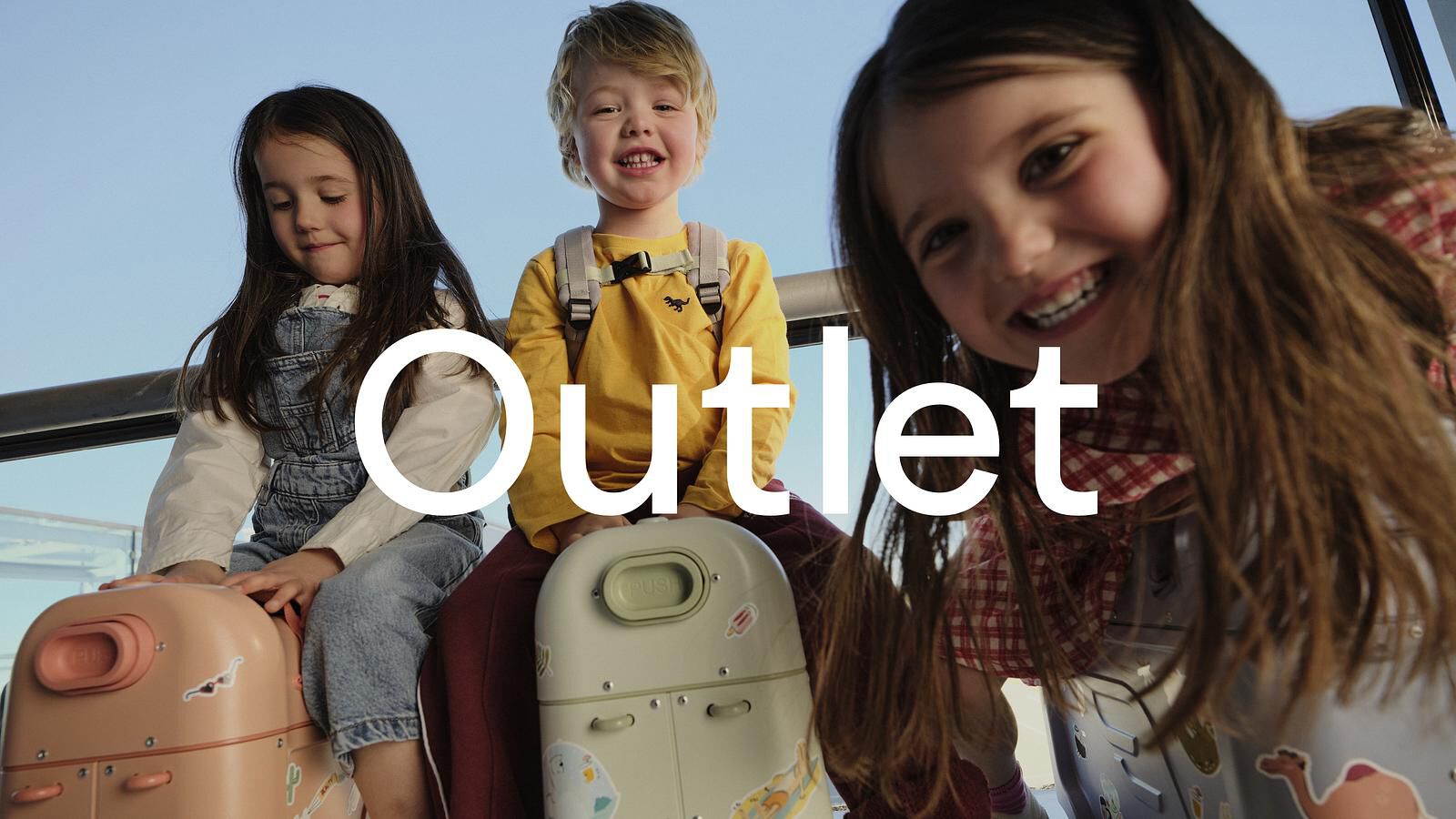 Trois enfants souriants sont assis sur des bagages pour enfants à roulettes sur fond de ciel bleu.