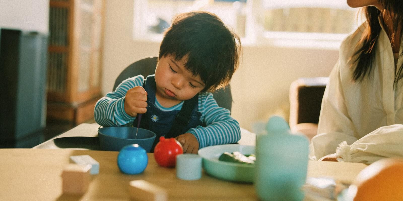 Un enfant concentré aux cheveux foncés est assis à une table, tenant un ustensile dans un bol bleu.