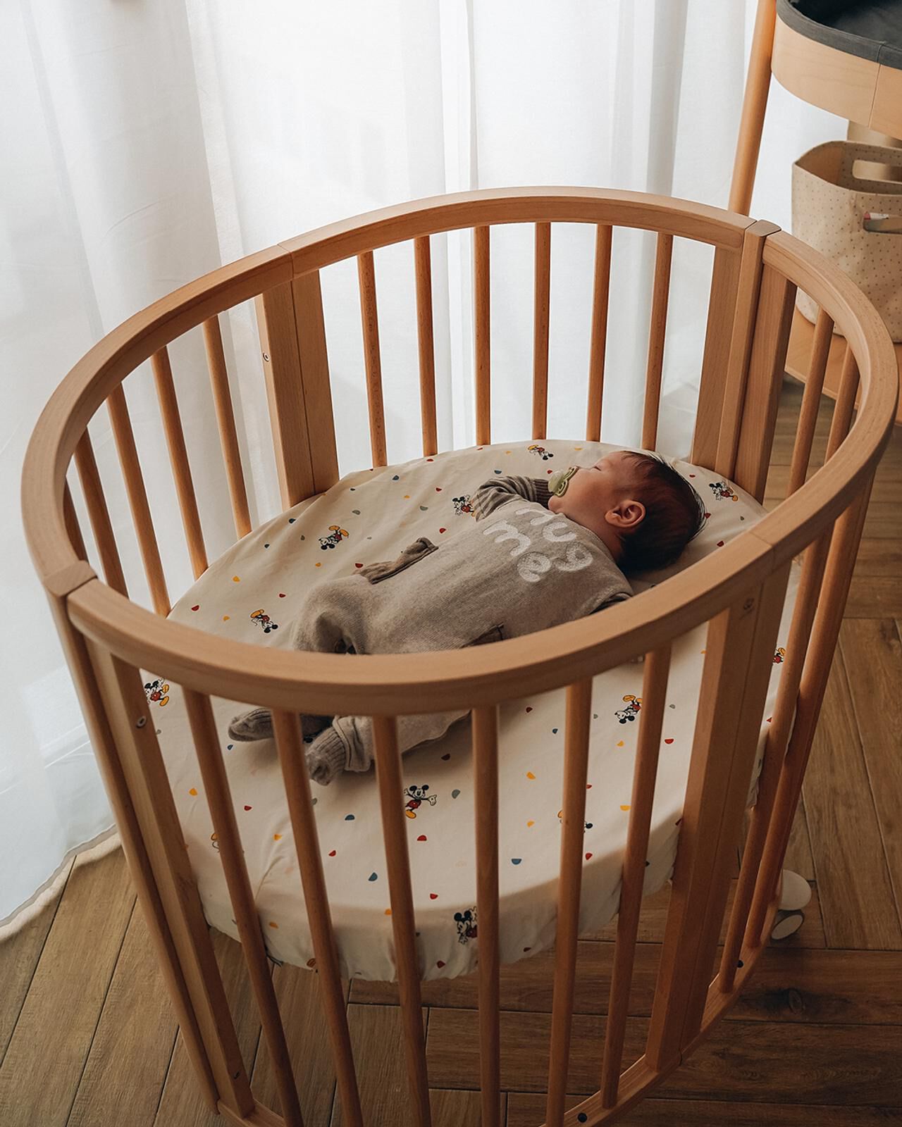 A baby sleeps soundly in an oval wooden crib on a Mickey Mouse sheet.