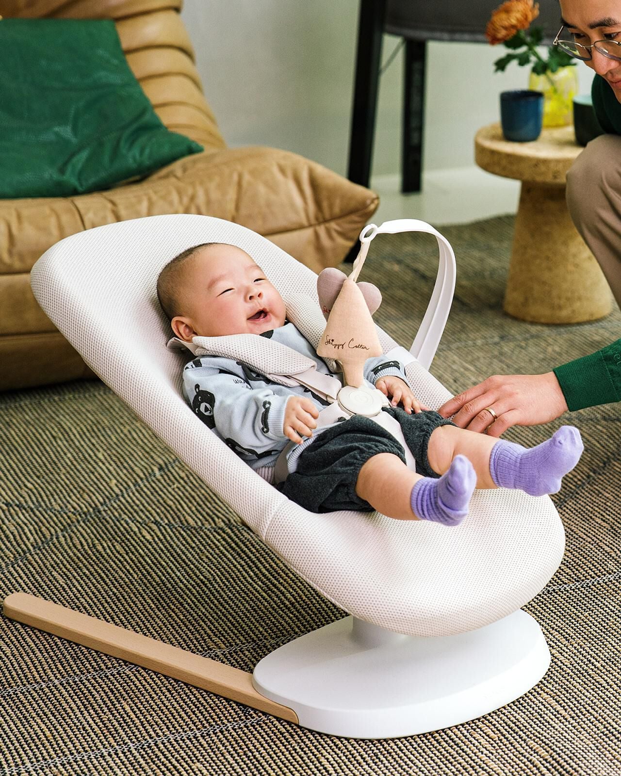 Smiling baby in a light bouncer with a wooden base, looking up at an adult's hands.