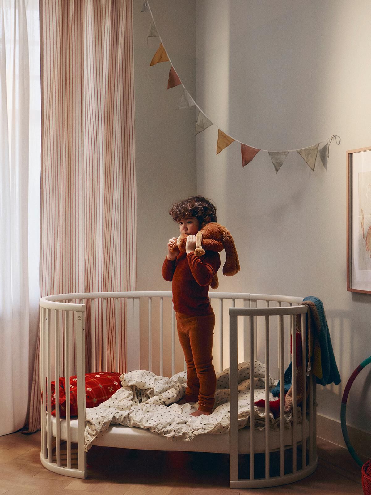 A toddler in rust pajamas stands in a Stokke Sleepi crib, hugging a brown stuffed bunny.