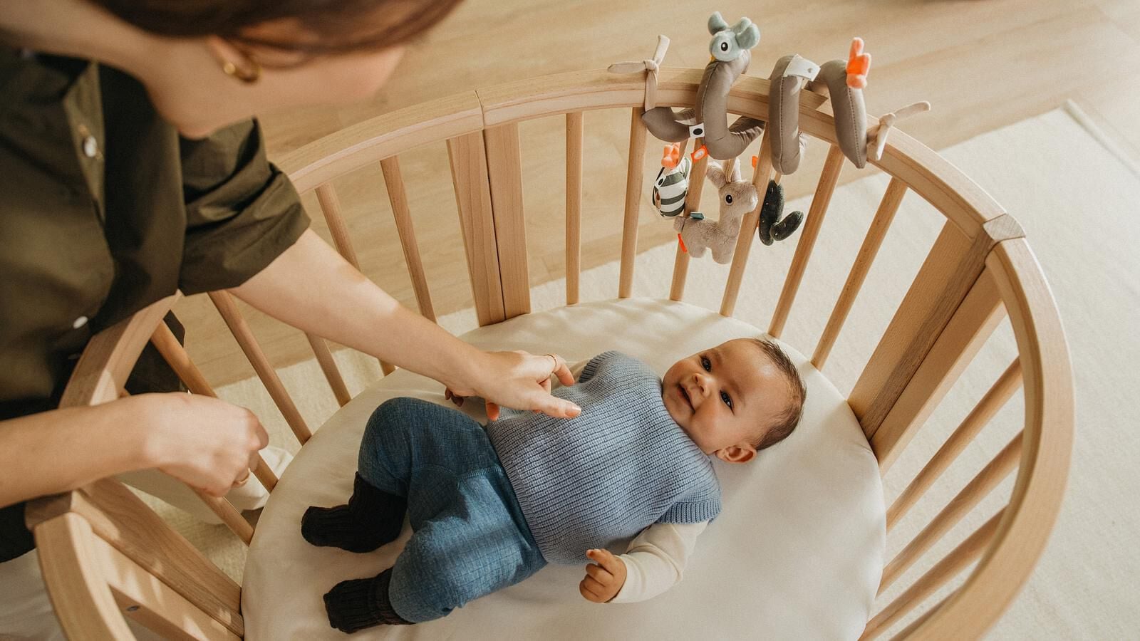 Een vrouw raakt voorzichtig een lachende baby aan die in een houten wieg met een spiraalvormig speeltje ligt.
