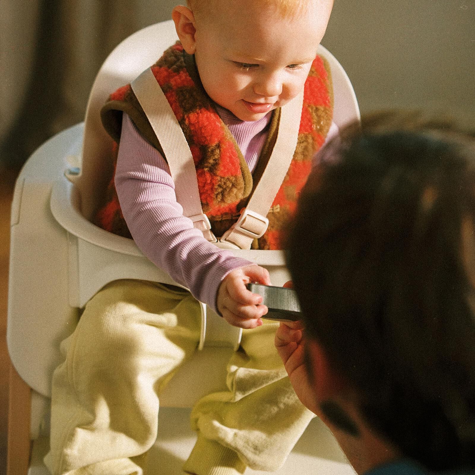 Un bambino in un seggiolone Tripp Trapp con Baby Set allunga la mano verso un oggetto tenuto da un adulto.