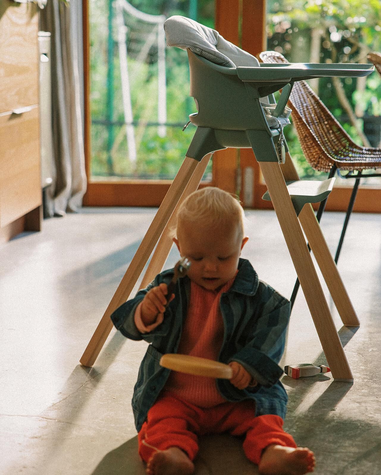 A baby sits playing with toys on the floor, with a modern high chair in the background.