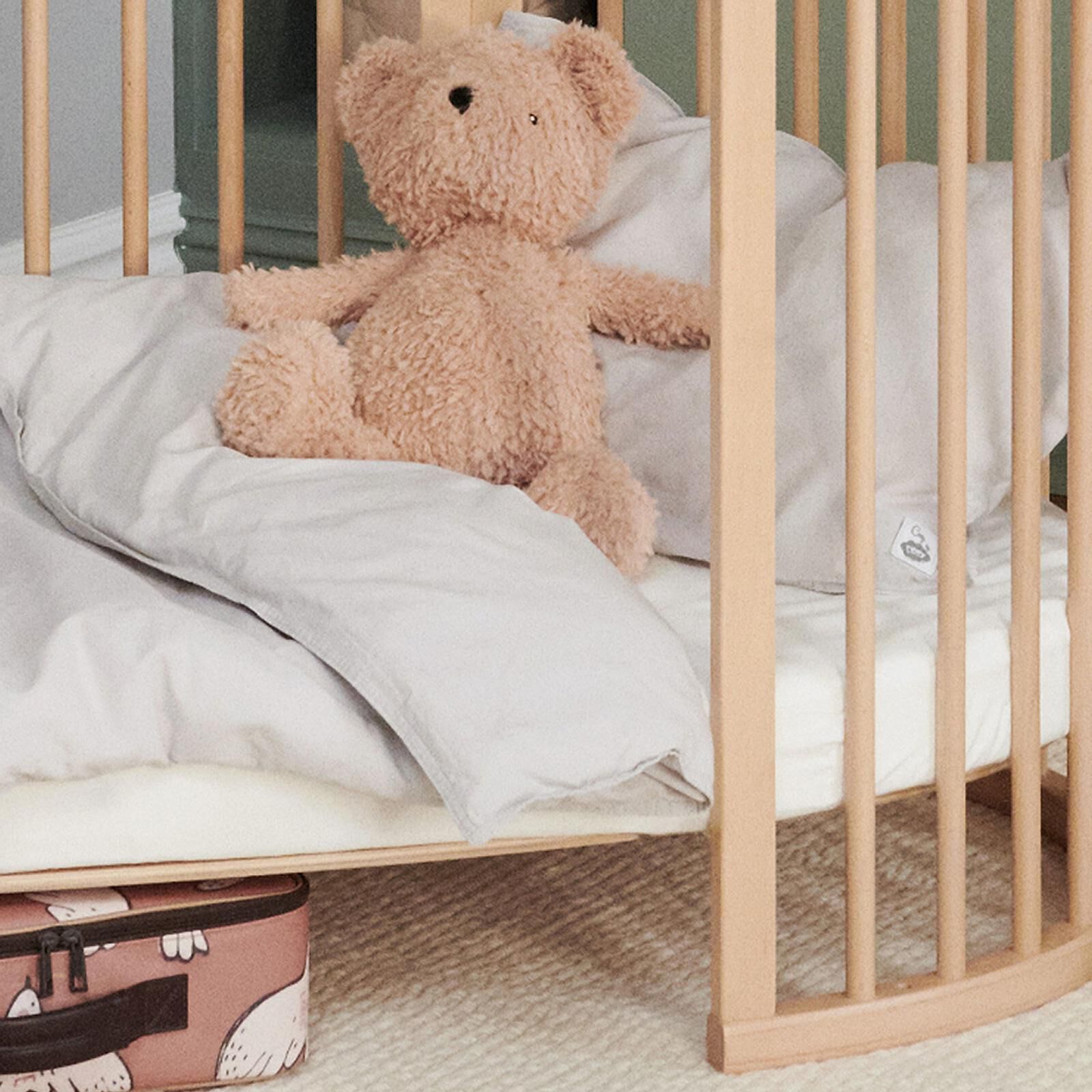 A light brown teddy bear sits upright in a wooden crib with light grey bedding. A pink suitcase is below.