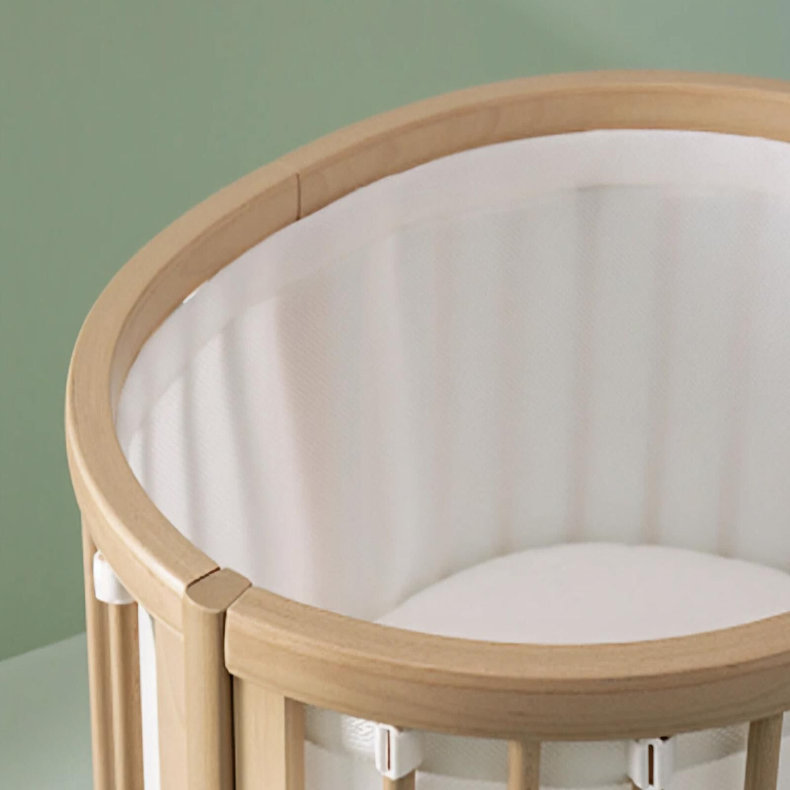 An oval wooden crib with white mesh lining and mattress against a green background.