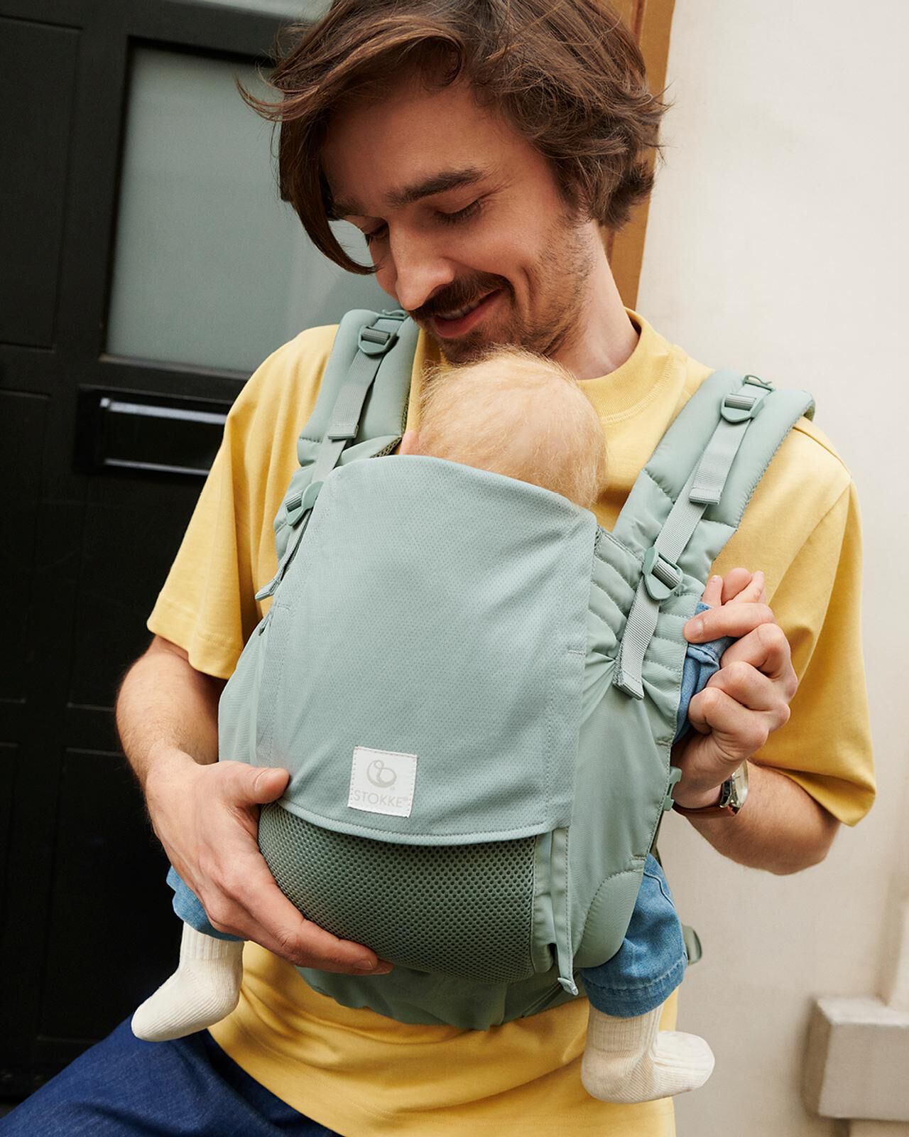 Happy dad carries baby in a light green baby carrier, looking down fondly.