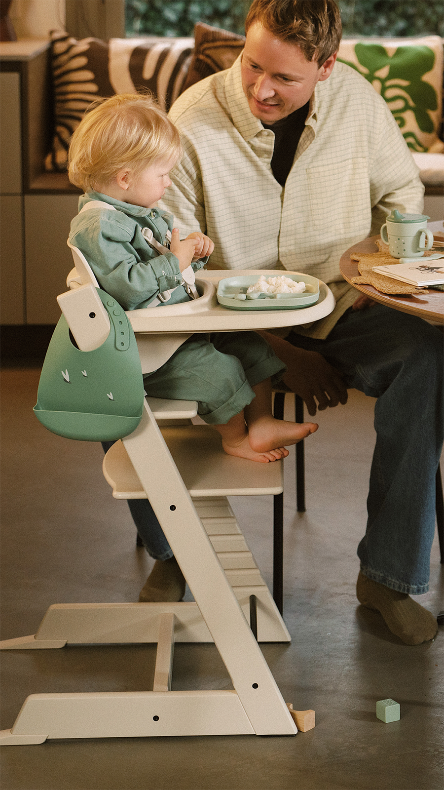 A man smiles at a child eating in a Tripp Trapp high chair with a green bib.