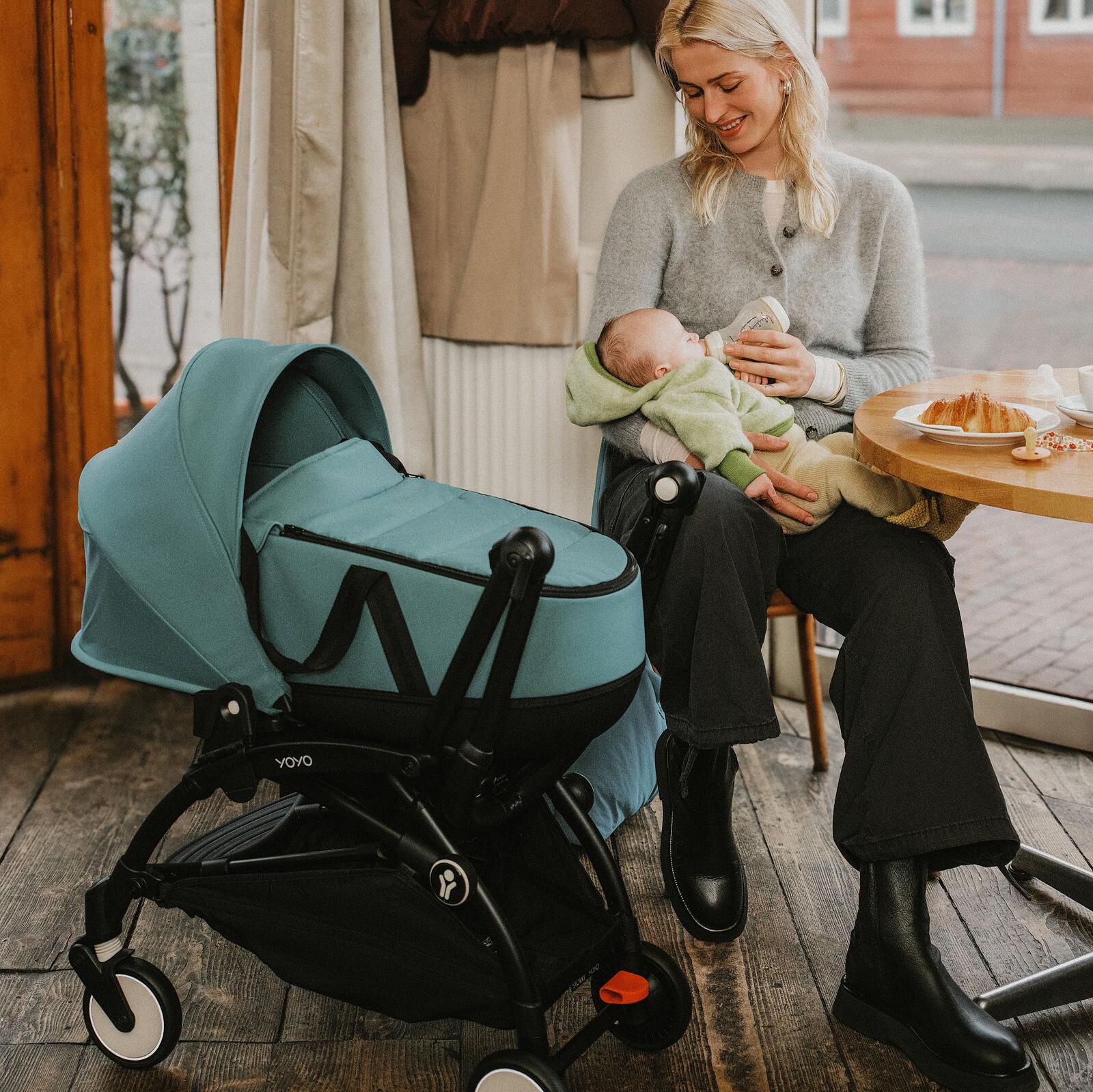 Eine Frau füttert ihr Baby mit der Flasche. Ein petrolfarbener STOKKE YOYO Kinderwagen steht in der Nähe in einem Café.