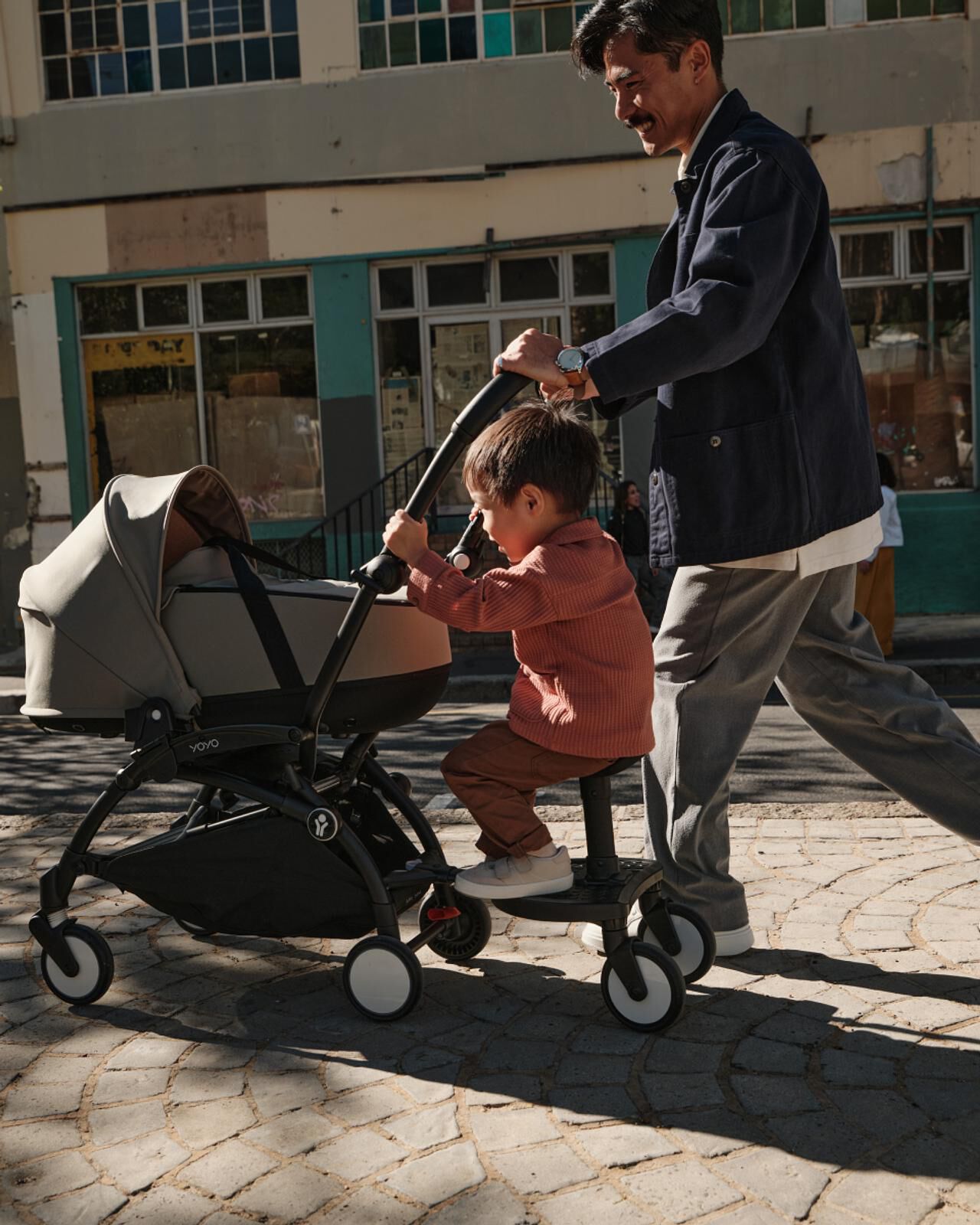 Lächelnder Mann schiebt einen STOKKE YOYO Kinderwagen mit einem Kind, das auf einem angebrachten Mitfahrbrett sitzt.