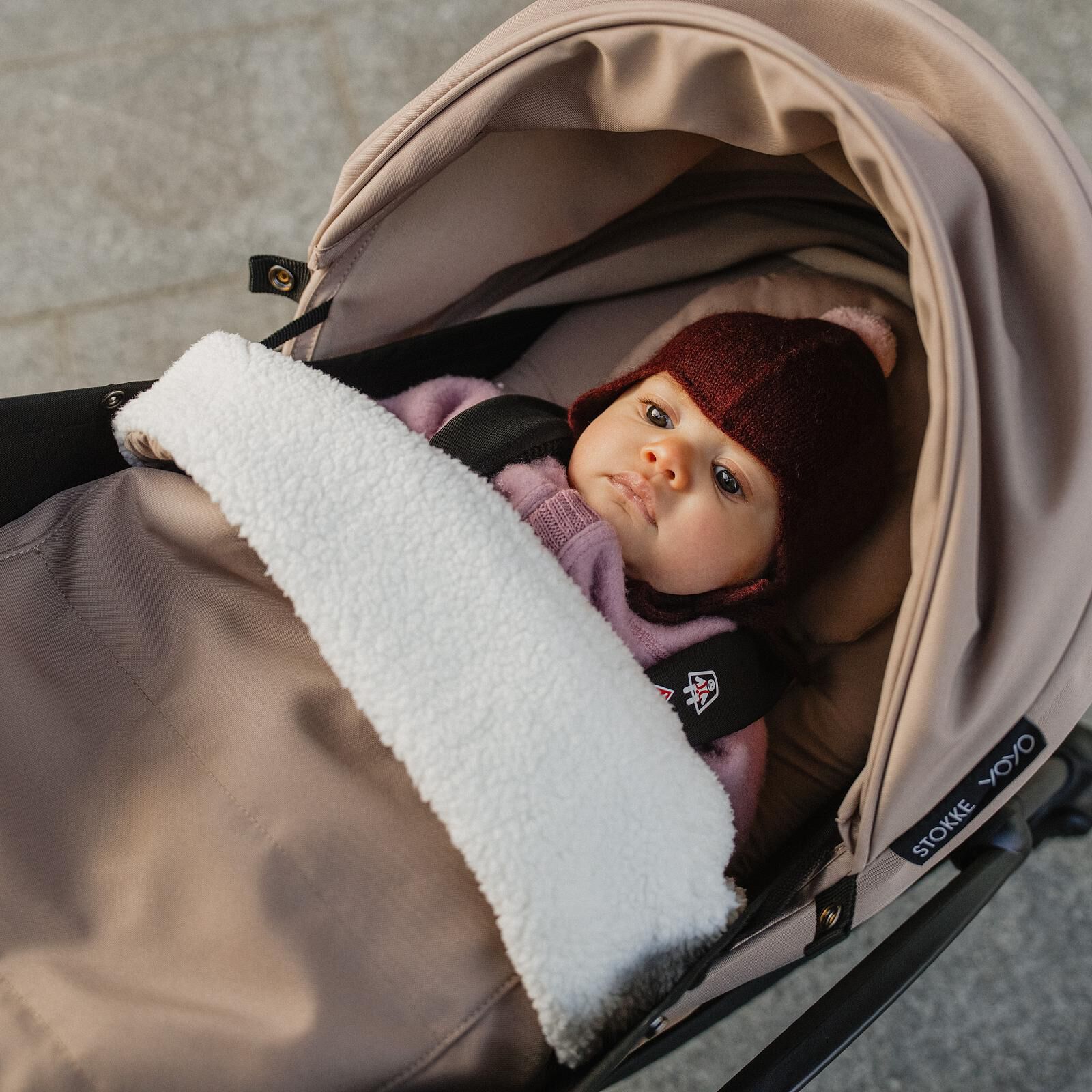 En baby med burgunderfarget lue og rosa klær, pakket inn i et hvitt teppe, i en beige STOKKE YOYO barnevogn.