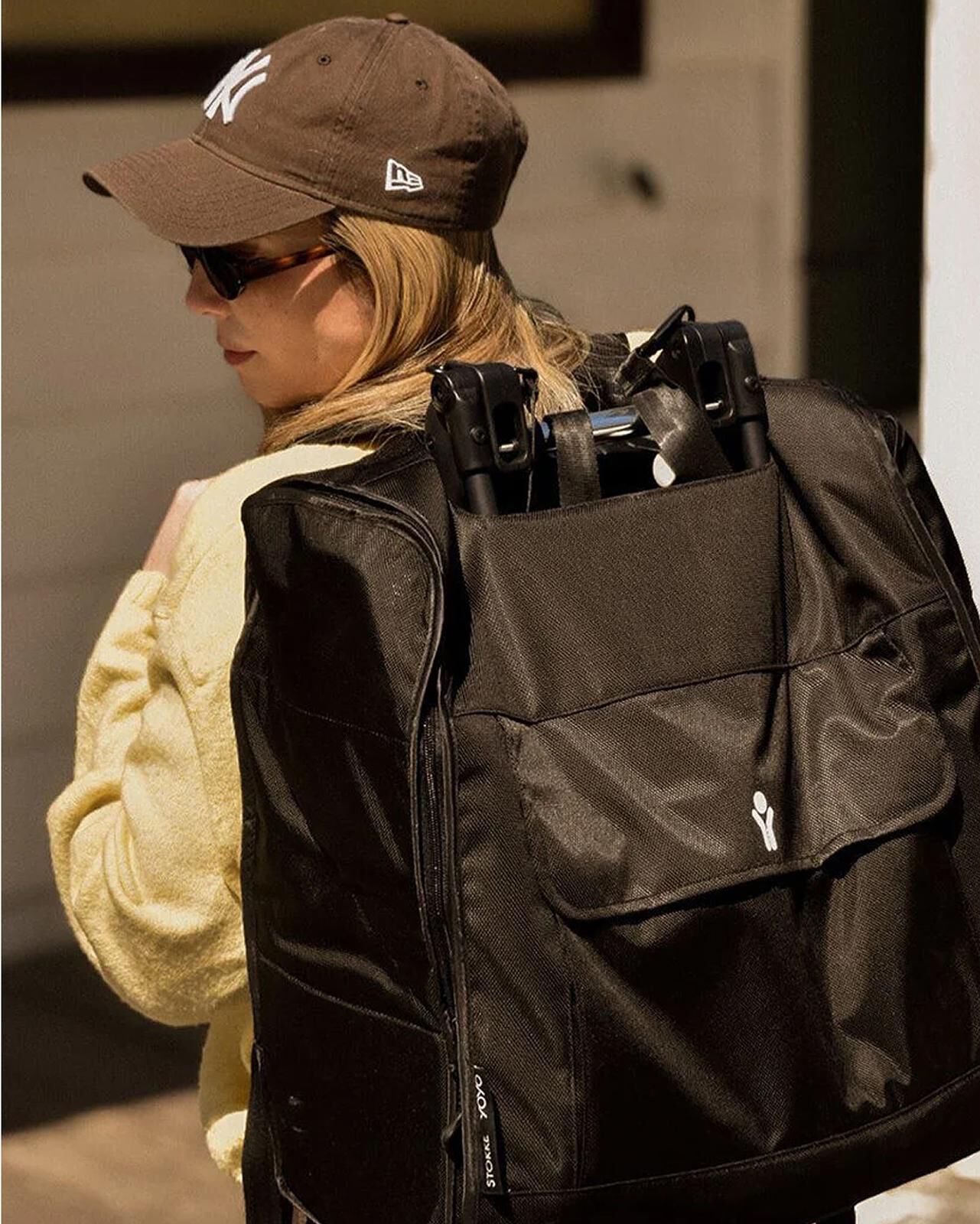 Blond woman in a Yankees cap carries a STOKKE YOYO stroller in a black travel bag.