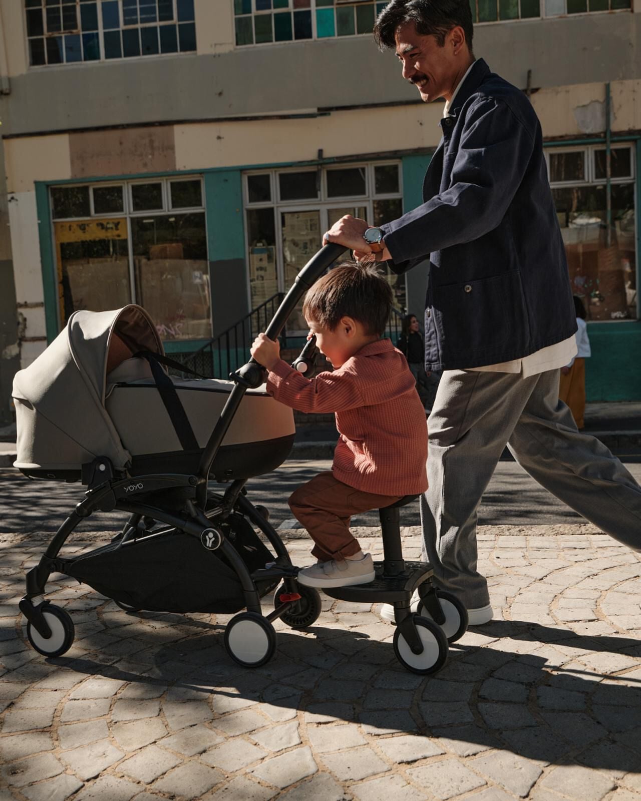 Un homme souriant pousse une poussette STOKKE YOYO avec un enfant assis sur une planche à roulettes attachée.