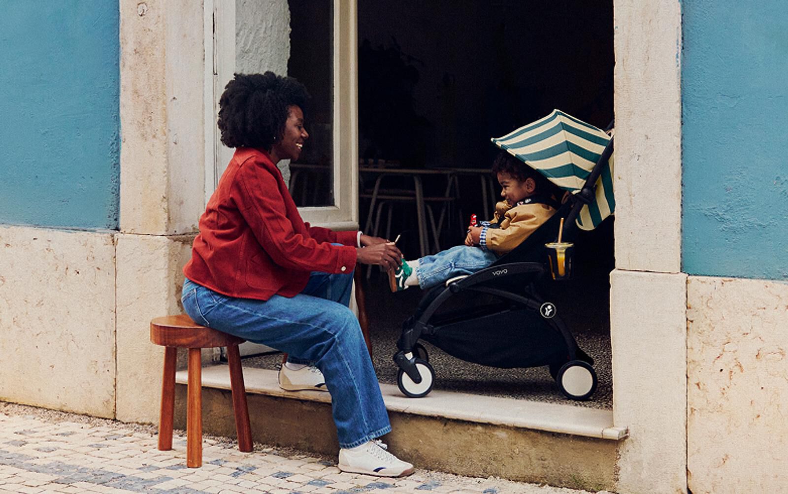 Eine lächelnde Frau bietet einem Kind in einem STOKKE YOYO Kinderwagen mit gestreiftem Sonnenschirm ein Getränk an.