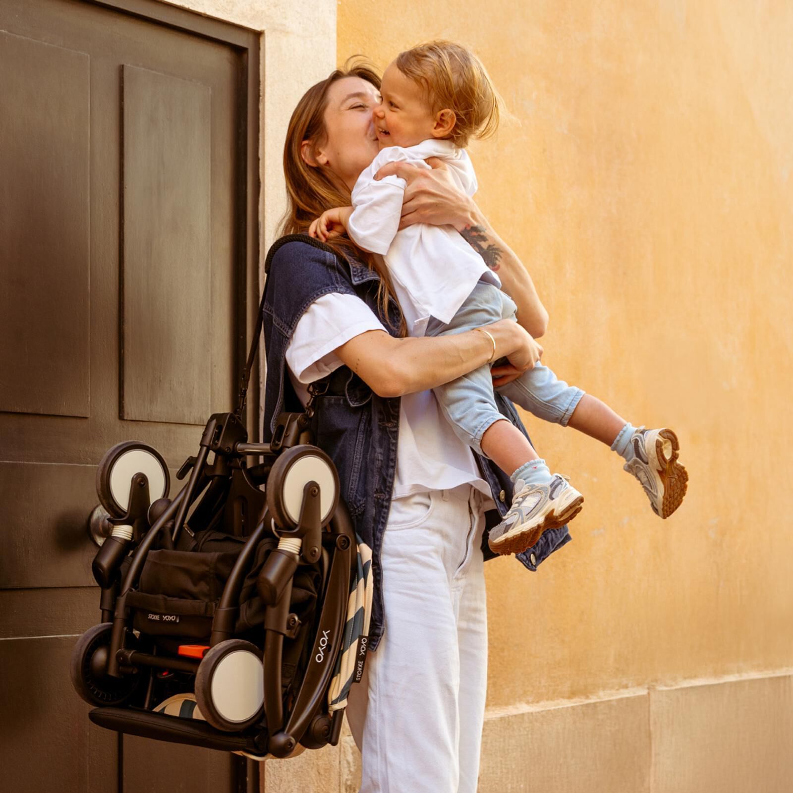 Mom kisses child, carrying a folded STOKKE YOYO stroller outdoors.