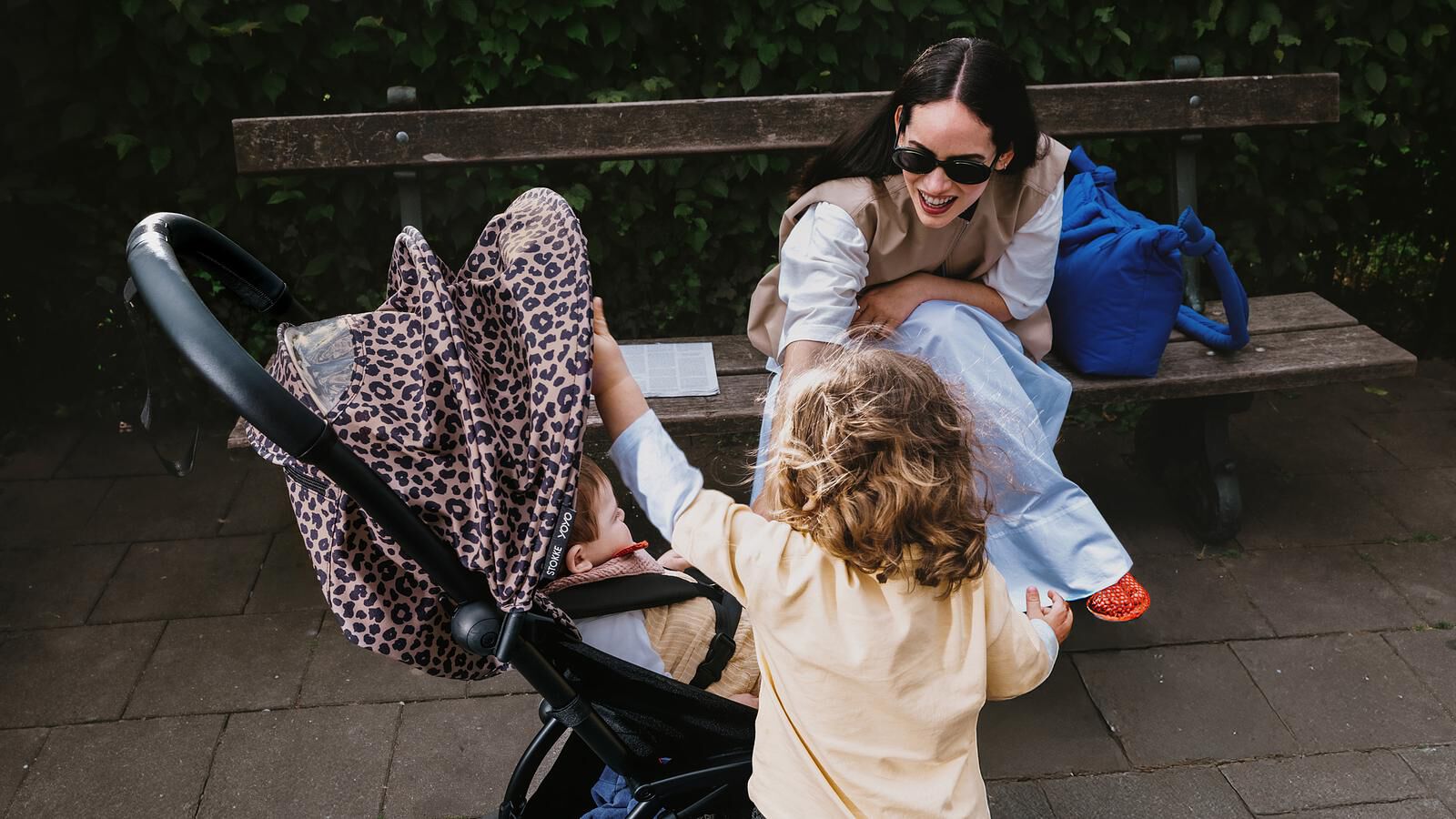 Eine lächelnde Frau auf einer Bank beobachtet, wie ein Kind nach einem Baby in einem STOKKE YOYO Kinderwagen mit Leopardenmuster greift.