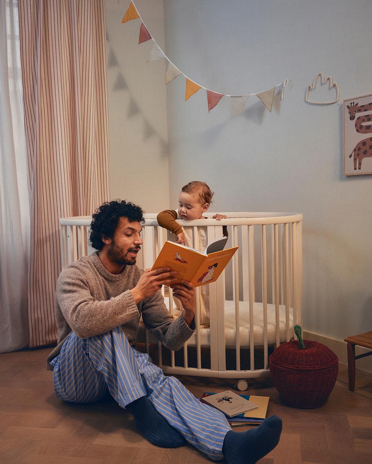 Father reads 'JANE'S BLANKET' to baby standing in a white crib.