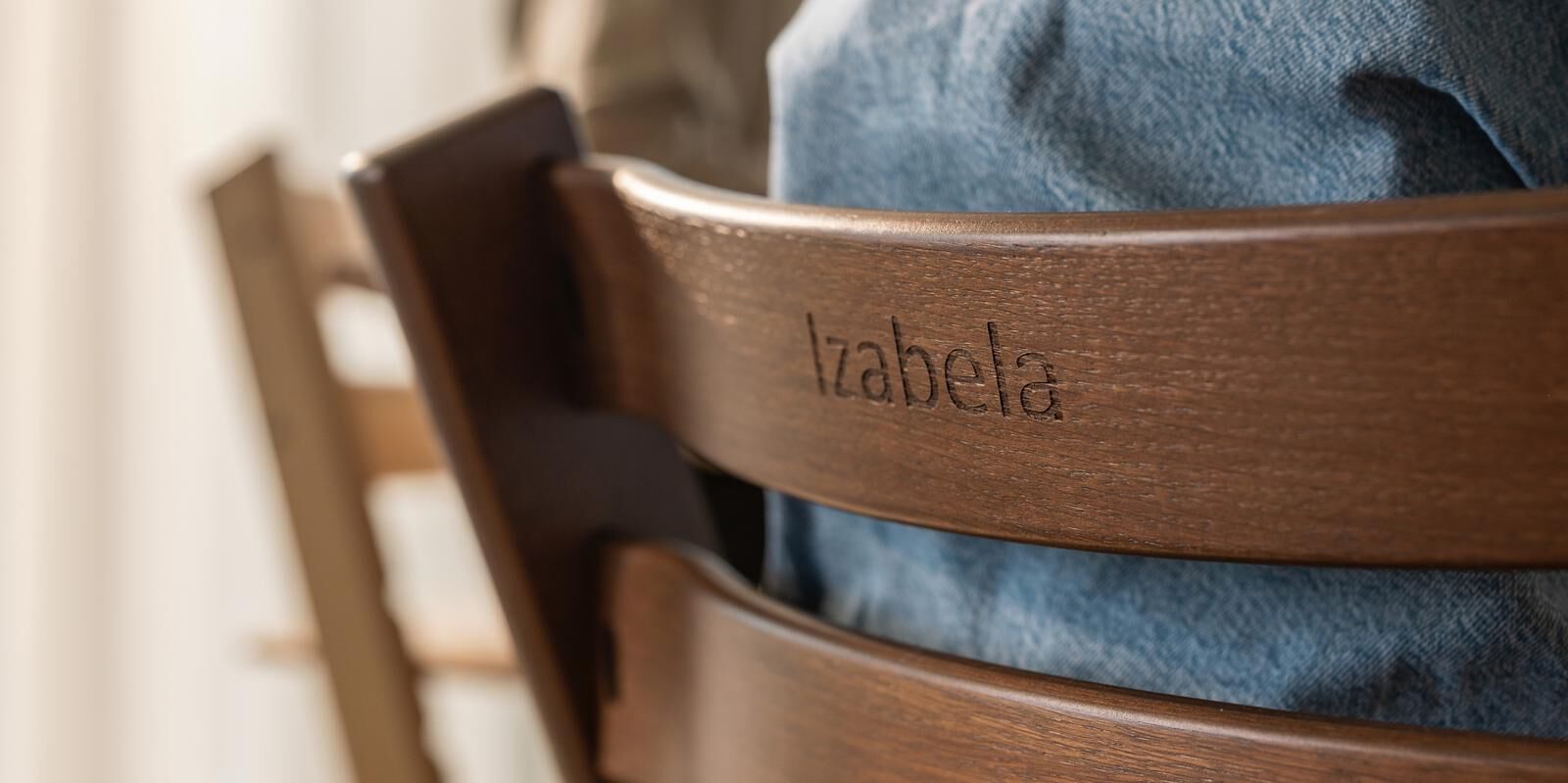 A person in blue denim sits in a dark wooden Tripp Trapp chair with 'Izabela' engraved.