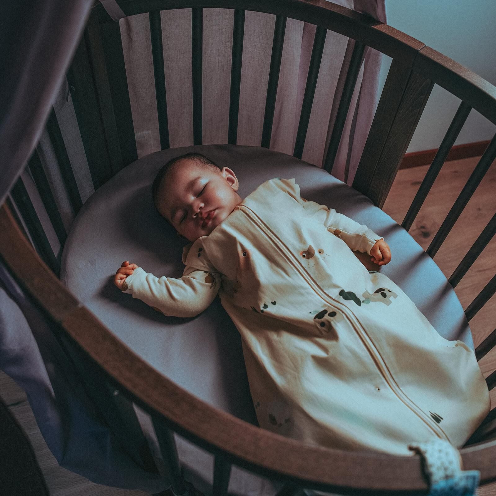 En baby i en mønstret sovepose sover fredelig i en oval treseng.