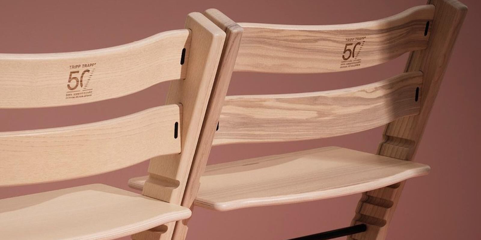 Two light wooden Tripp Trapp 50th Anniversary chairs against a reddish background.