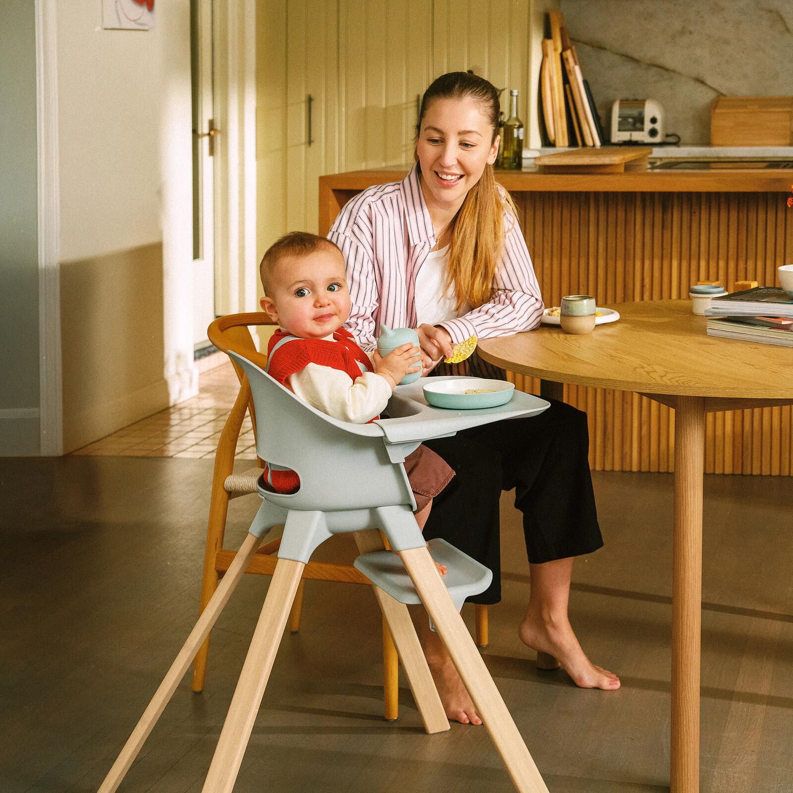 A smiling baby sits in a grey high chair, holding a cup, with a smiling woman at a wooden table.