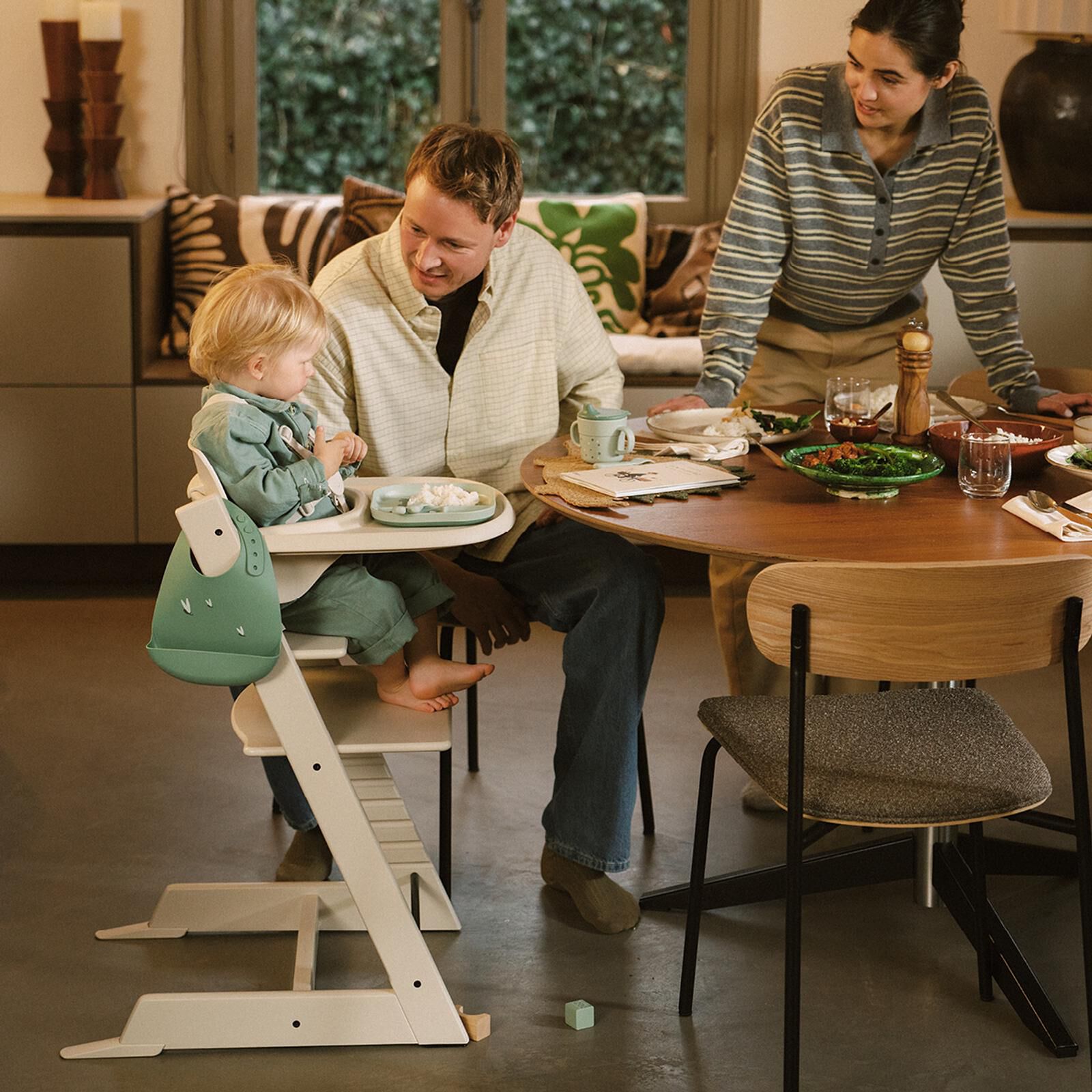 A family shares a meal. A toddler sits in a Tripp Trapp high chair with a green bib.