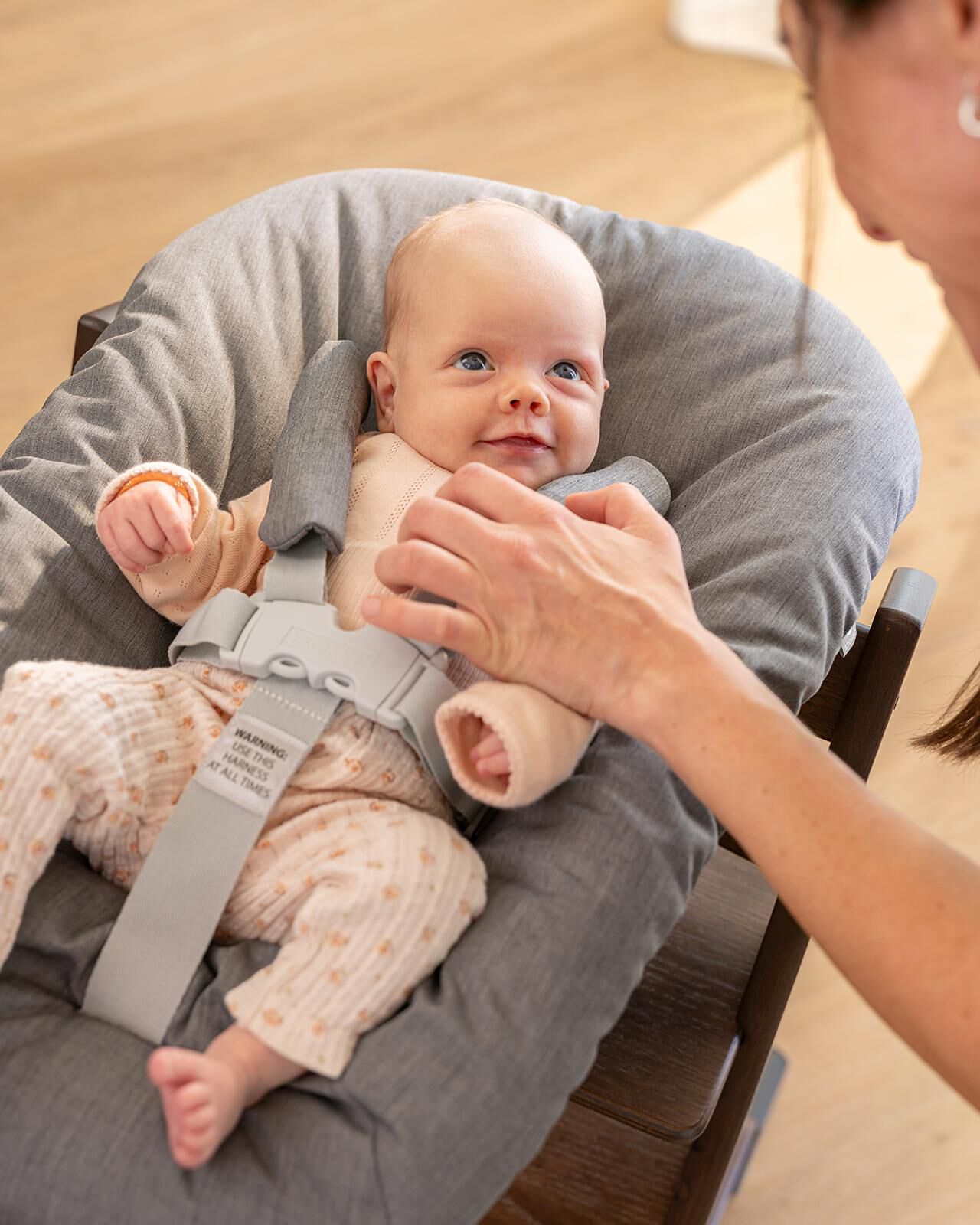Smiling baby in a Tripp Trapp high chair, a woman's hands adjusting the harness.