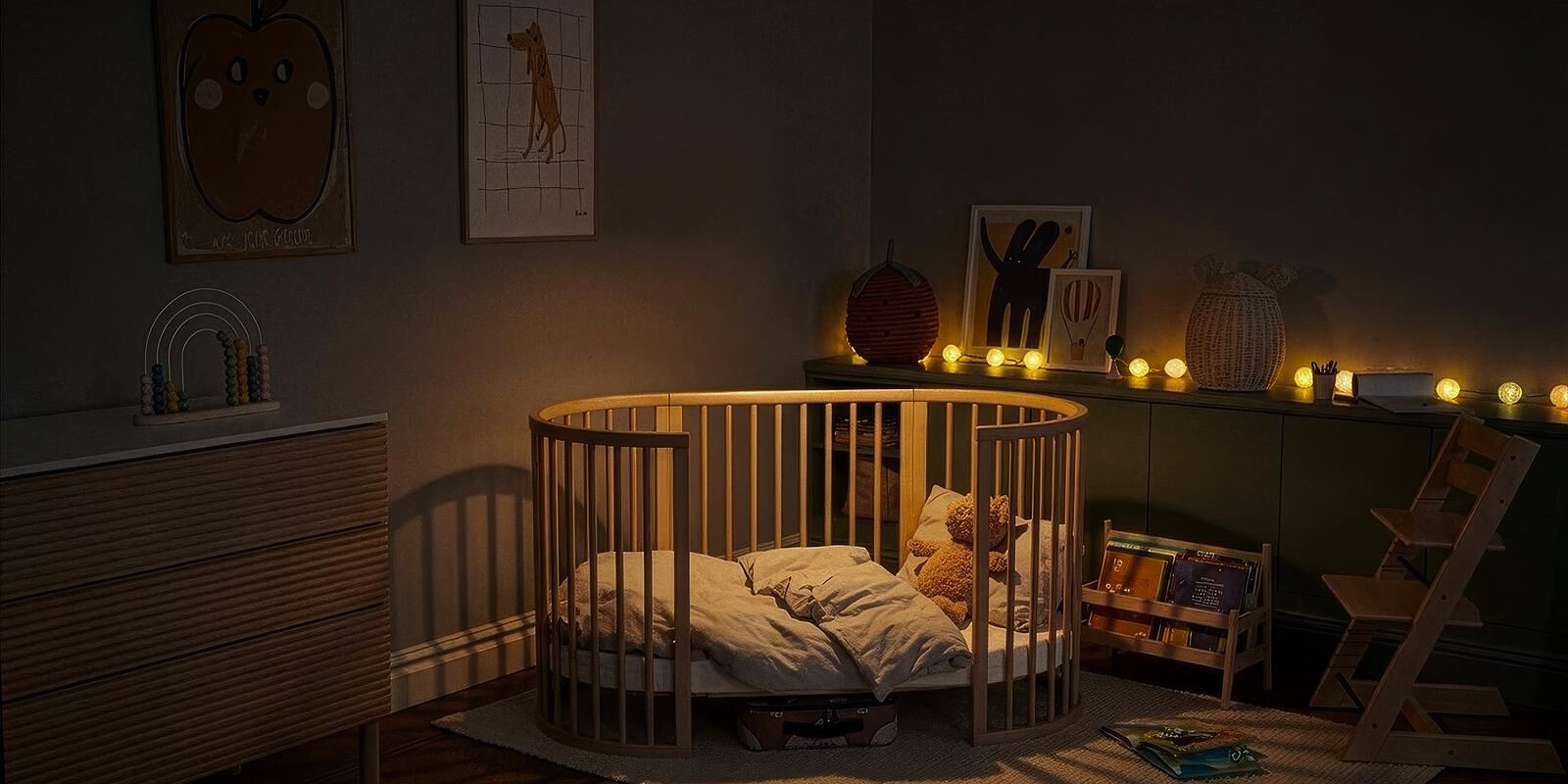 A cozy, dimly lit nursery with a round wooden crib, a teddy bear, and a Tripp Trapp chair.