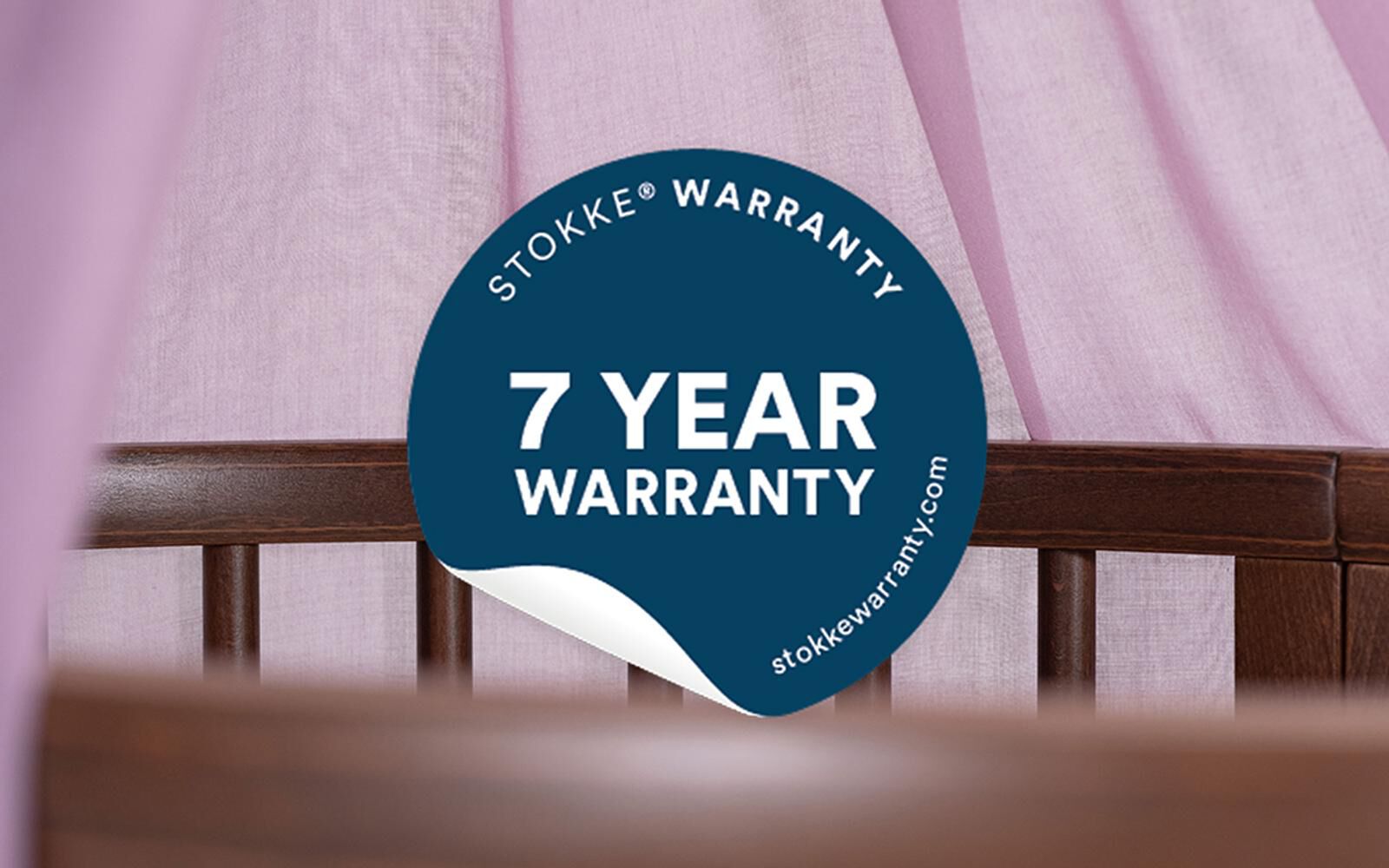 Blue Stokke 7 Year Warranty sticker on a wooden crib, with a pink curtain in the background.
