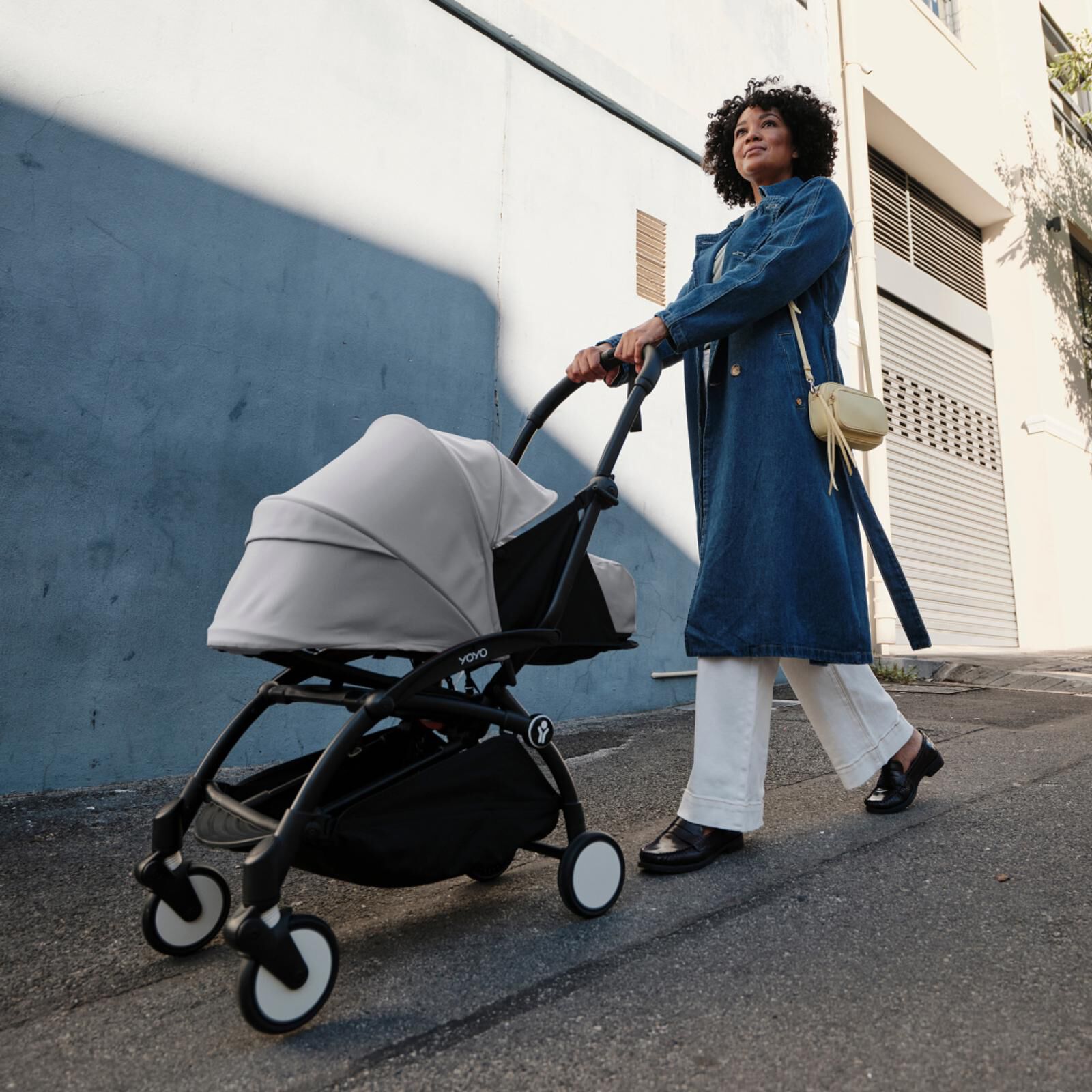 Femme en manteau en jean poussant une poussette STOKKE YOYO avec un auvent gris dans une rue urbaine.