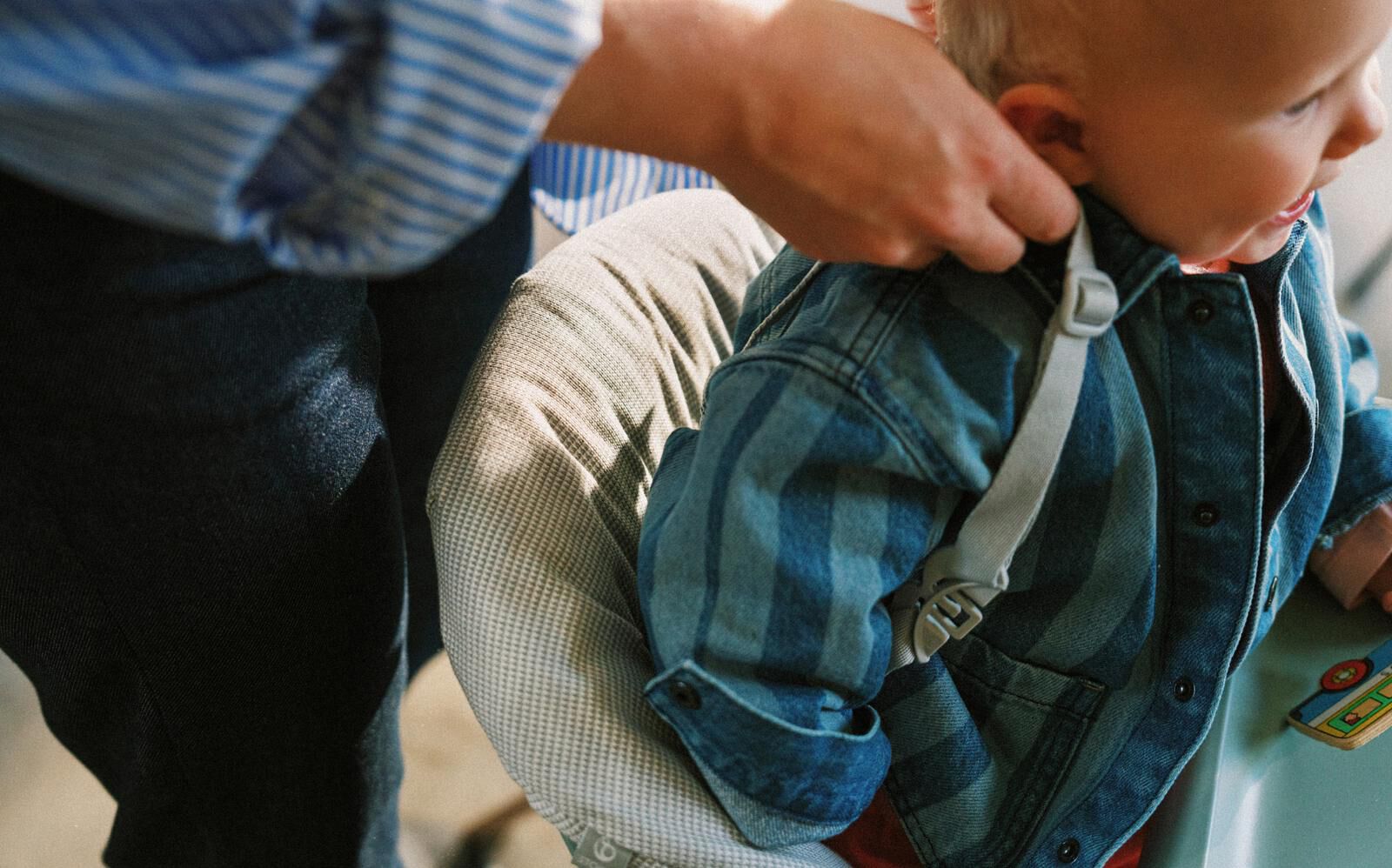 Adult fastens harness on baby in soft baby seat. Baby wears blue striped denim jacket.