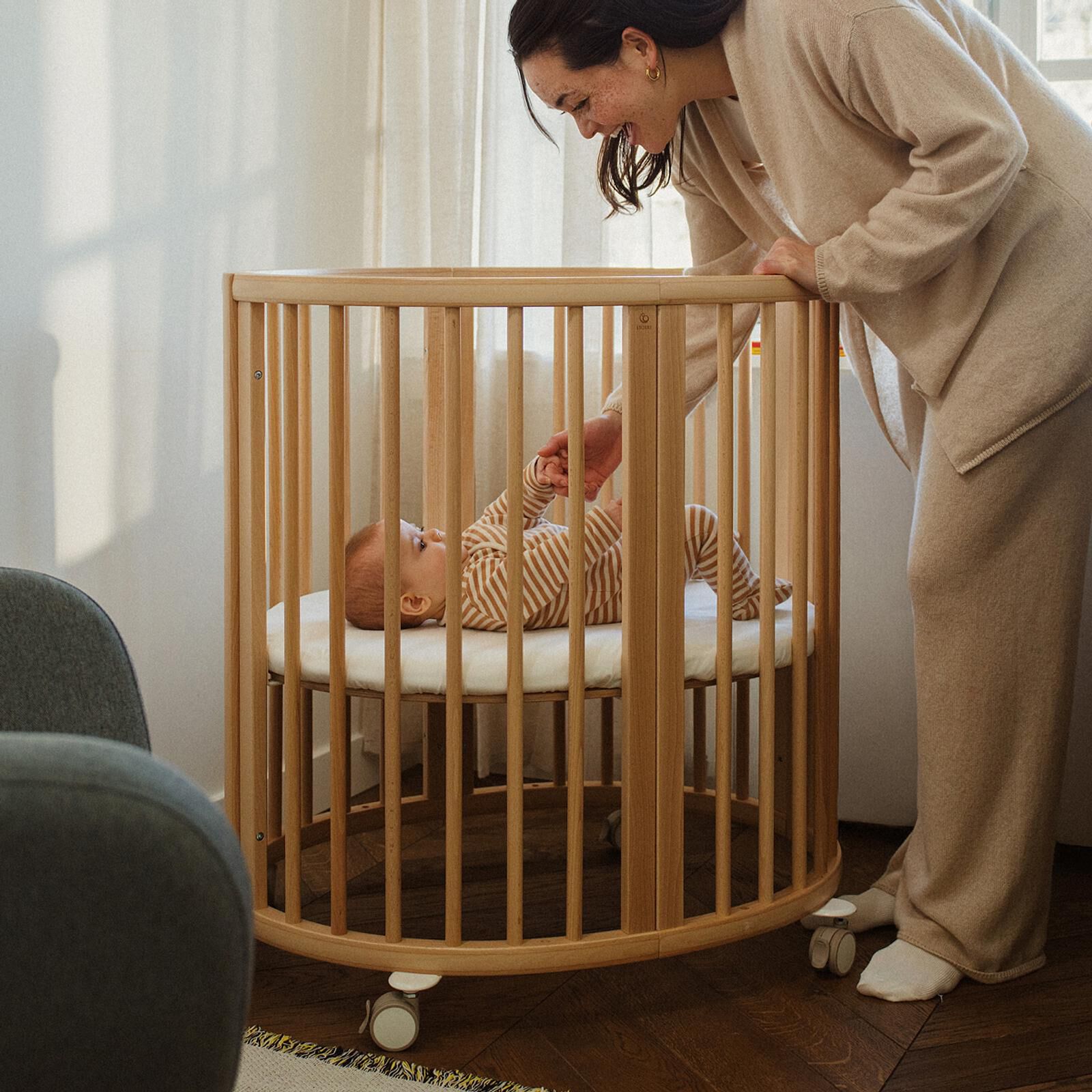 Smiling mother holds baby's hand in a round wooden crib.