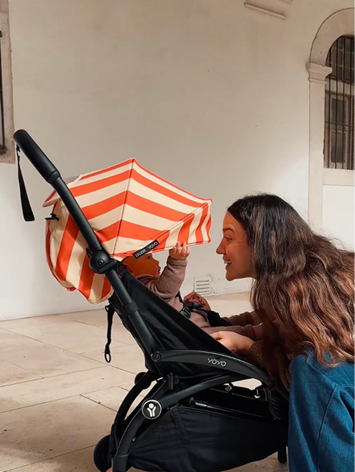 Eine lächelnde Frau beugt sich über einen YOYO-Kinderwagen mit einem orange-weiß gestreiften Sonnenschirm und schaut ein Baby an.