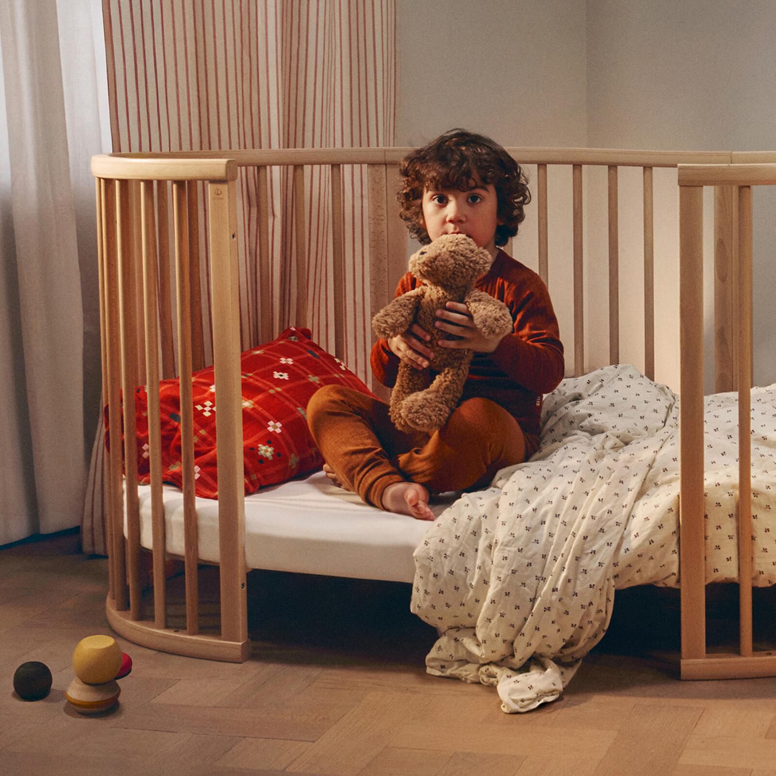 Child sits in an oval wooden crib, holding a brown teddy bear.
