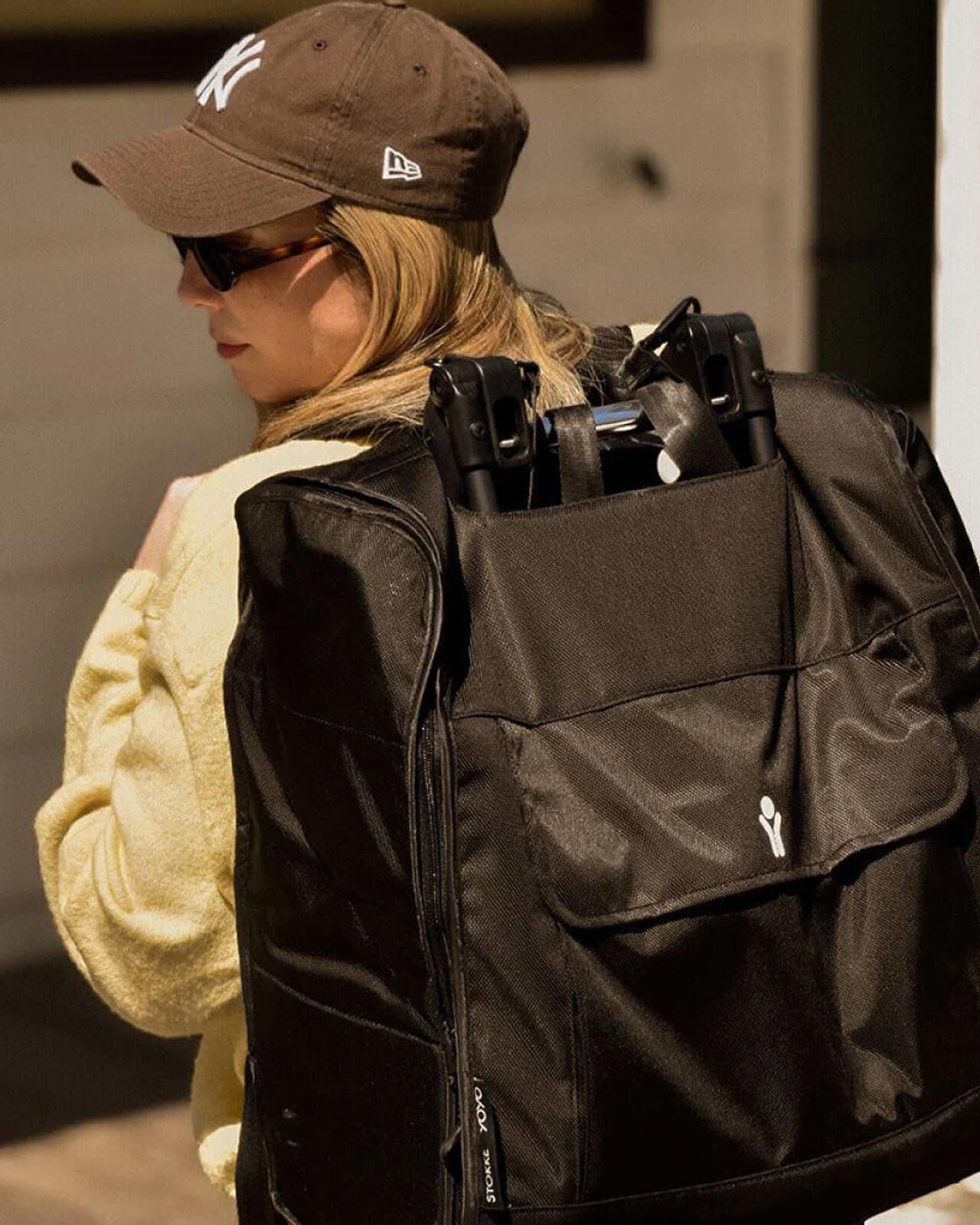 A woman wearing a Yankees cap and sunglasses carries a STOKKE YOYO stroller in a black travel bag.