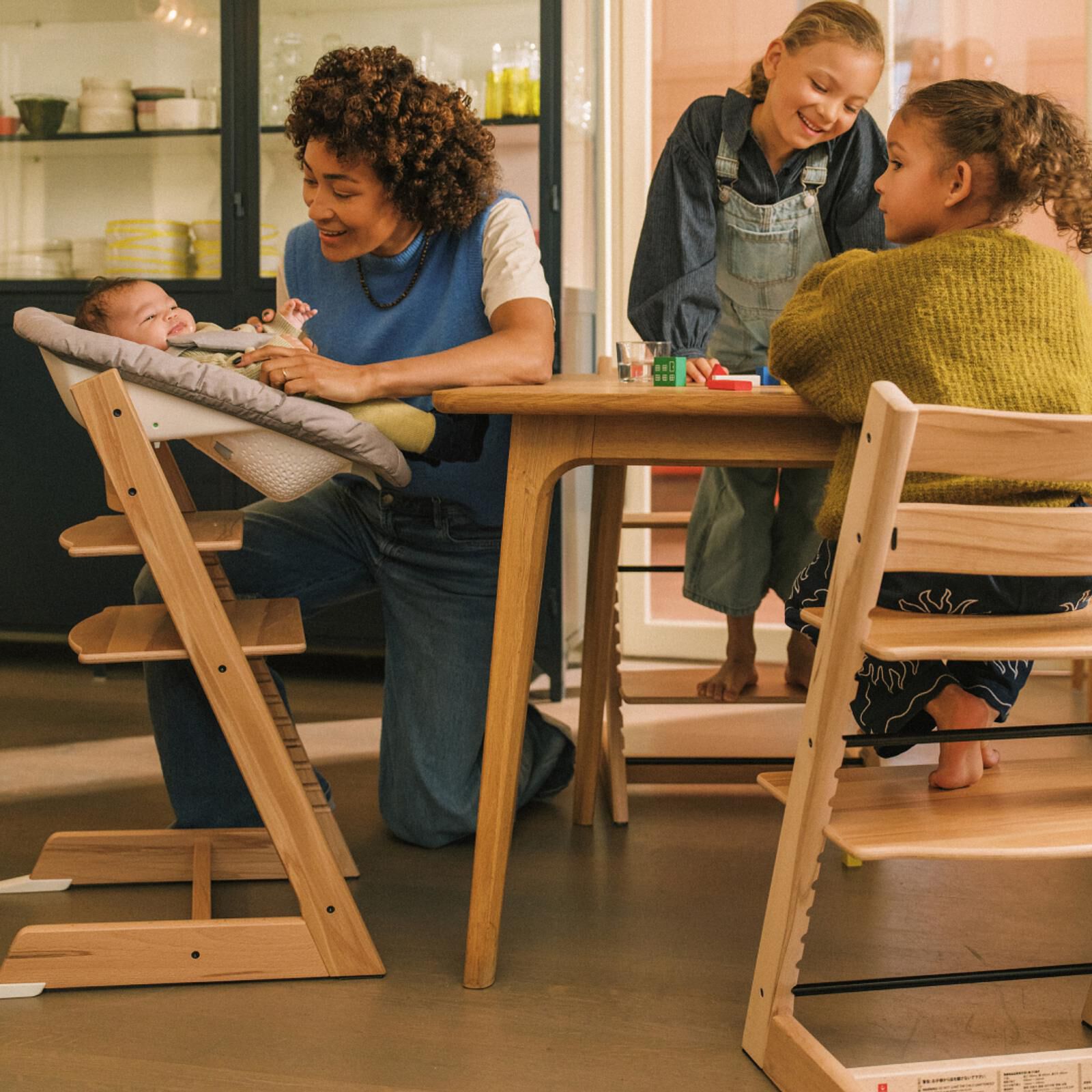 Vrouw lacht naar baby in Tripp Trapp kinderstoel, twee kinderen spelen aan een houten tafel met nog een Tripp Trapp.