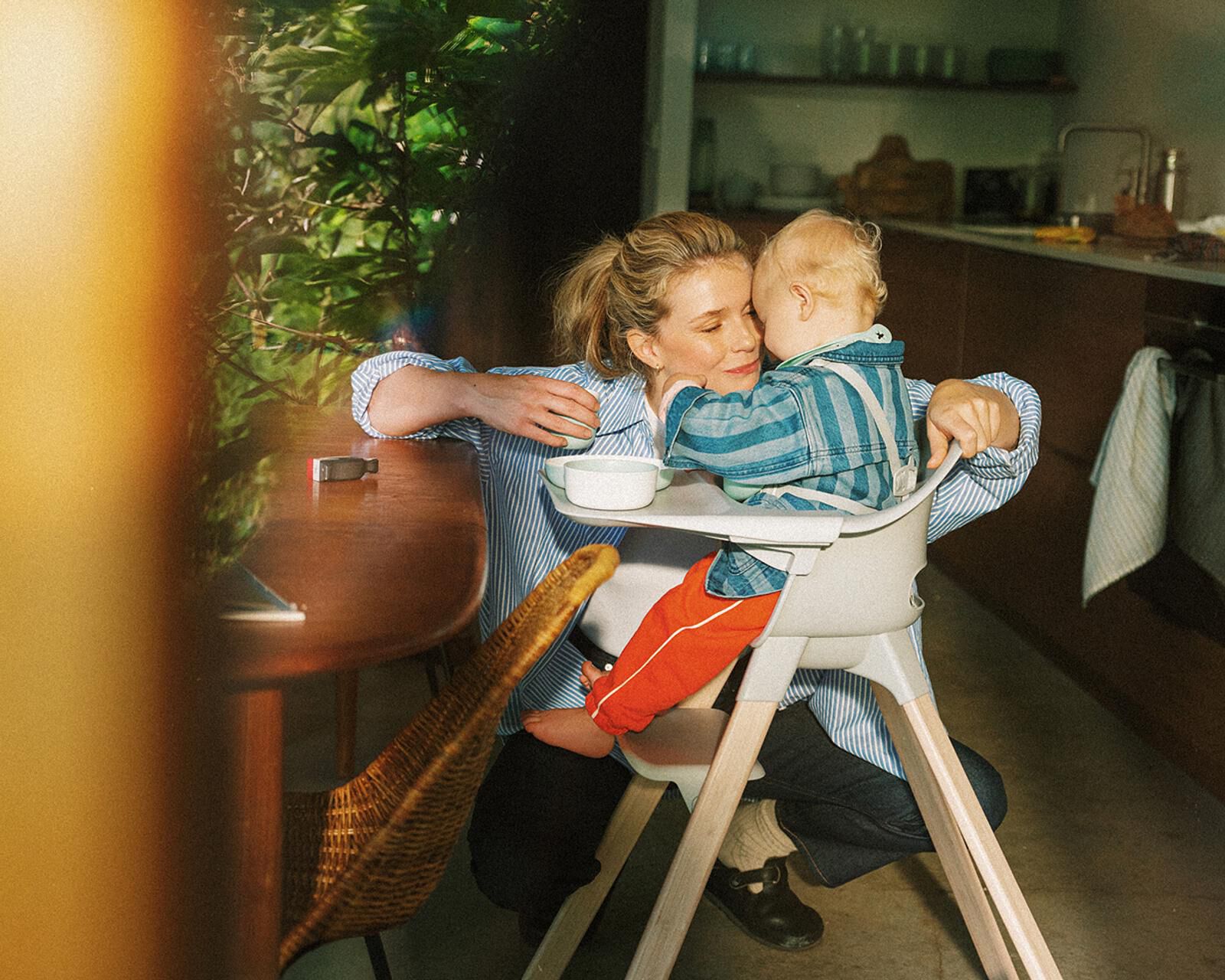 A mother lovingly embraces her baby sitting in a grey high chair at a wooden table.