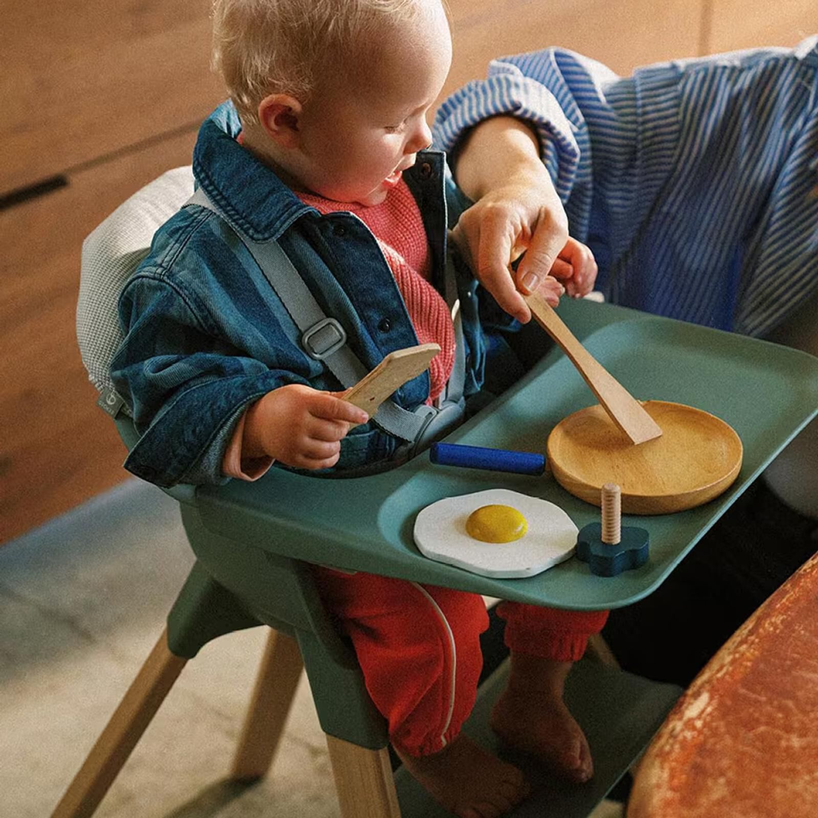 Child in a green high chair plays with toy fried egg and wooden spatula with an adult.