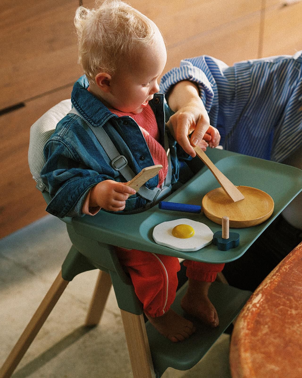 A baby plays with toy food on a tray in a green high chair, with an adult's hand assisting.