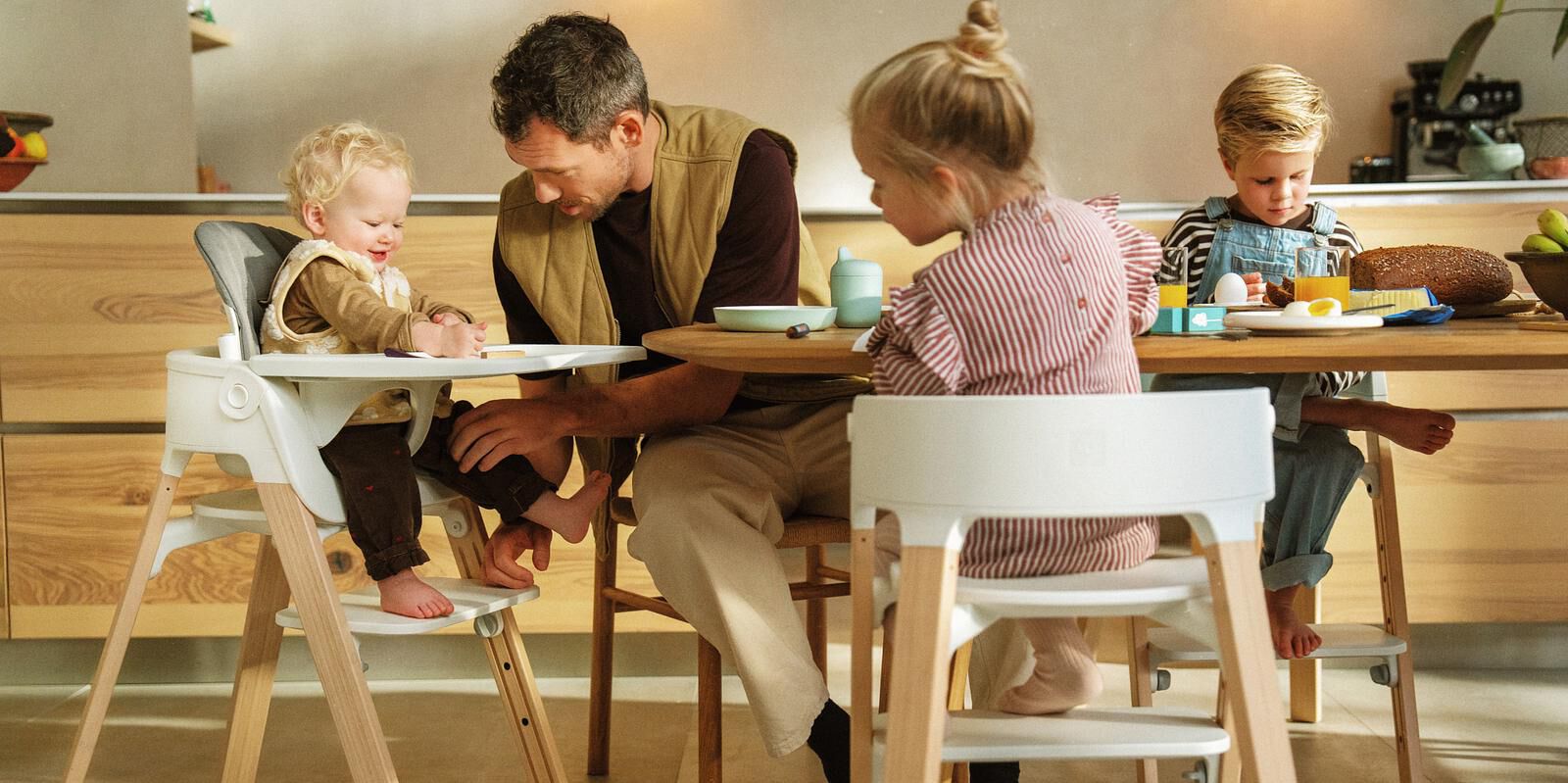 Un père aide un bébé souriant assis dans une chaise haute blanche à une table avec deux autres enfants.