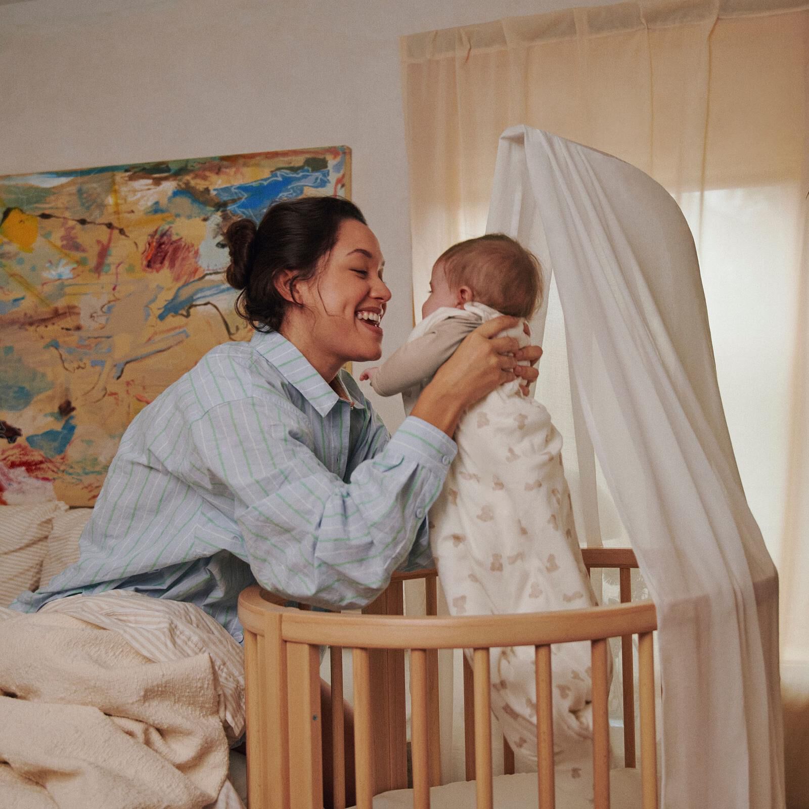Happy mother plays with her baby standing in a wooden crib with a canopy.