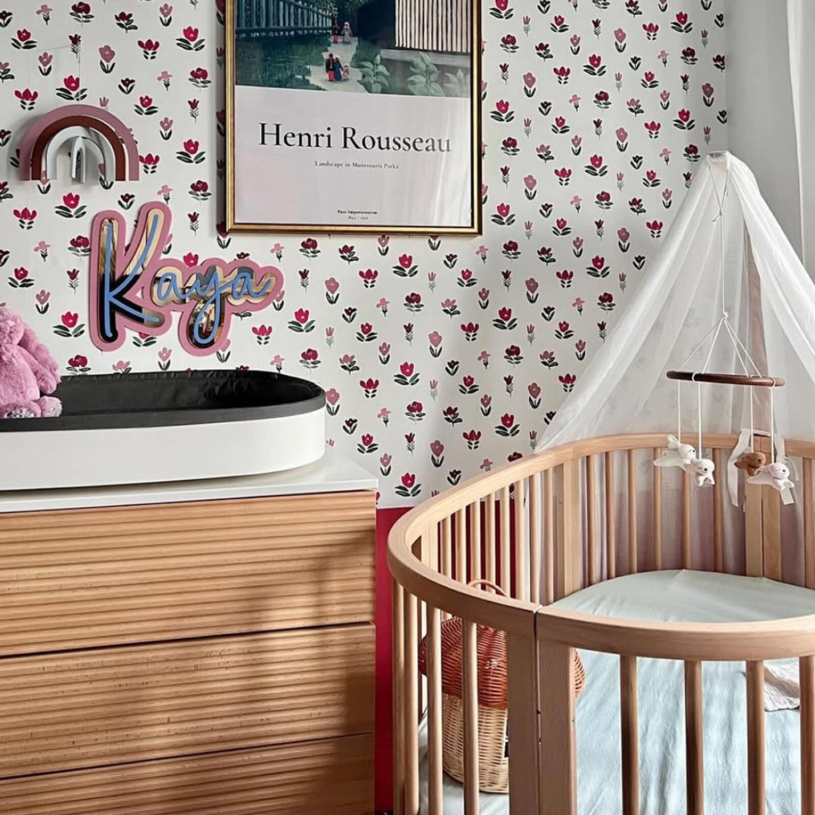 A cozy nursery with a wooden crib, changing station, floral wallpaper, and a 'Kaya' name sign.