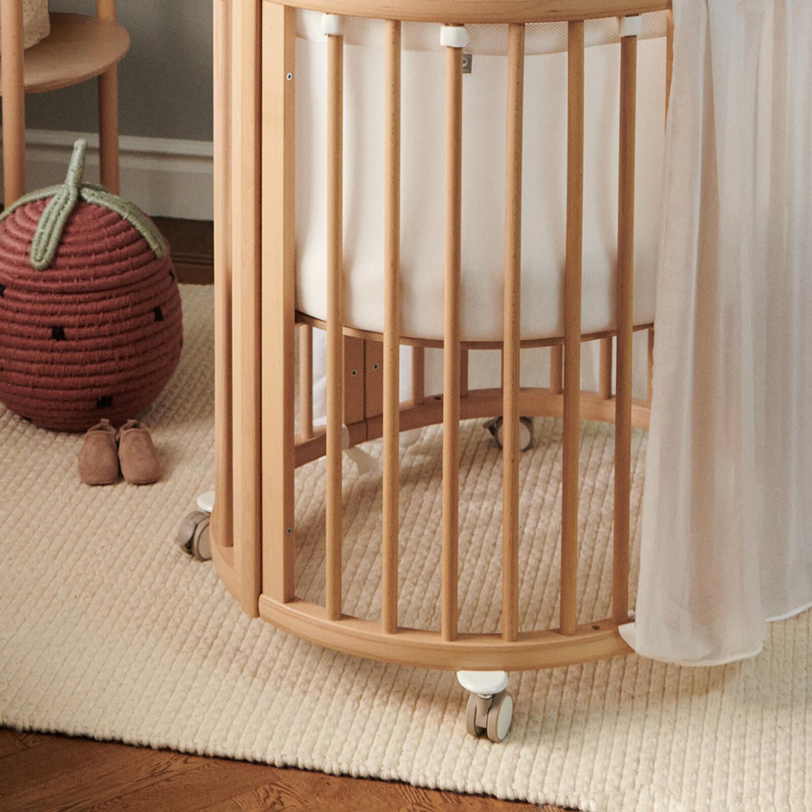 Un berceau ovale en bois avec de la literie blanche, de petites chaussures de bébé et un panier de fraises sur un tapis.