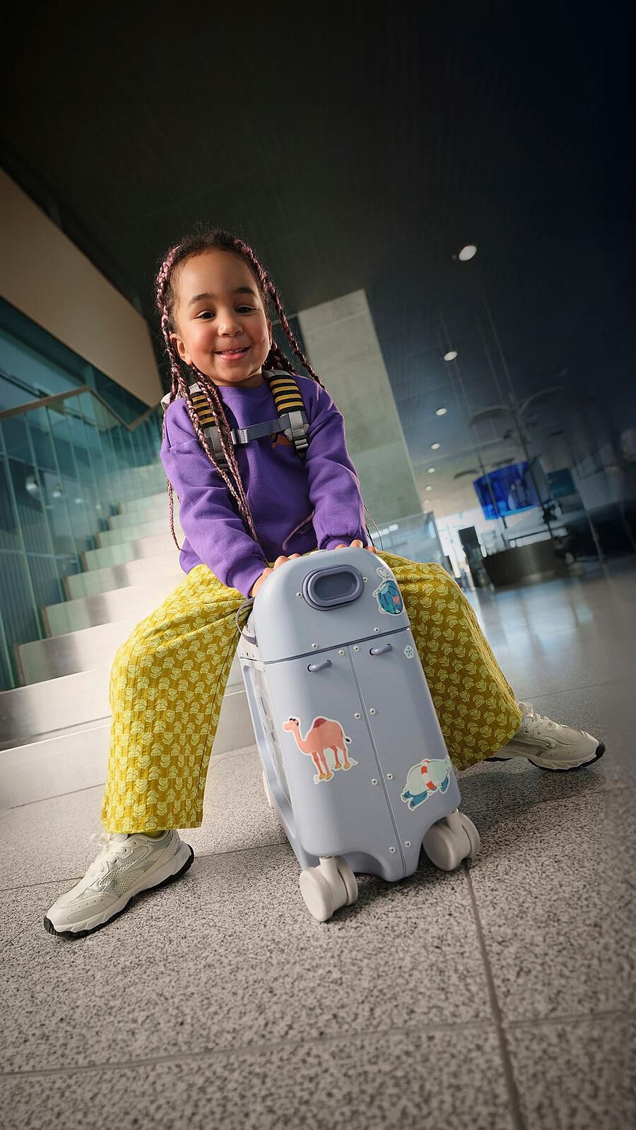Ein lächelndes Kind mit Zöpfen sitzt auf einem blauen Aufsitzkoffer mit Tieraufklebern in einem Flughafen.