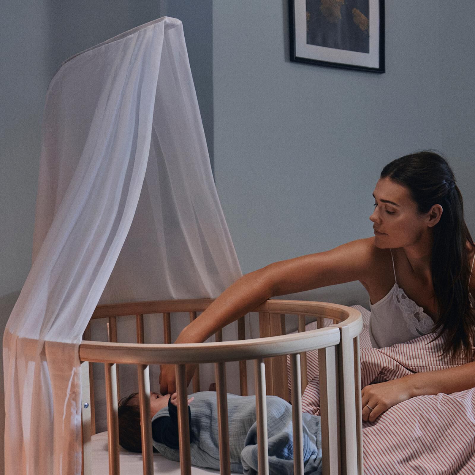 A mother gently tends to her baby sleeping in a wooden oval crib with a canopy at night.