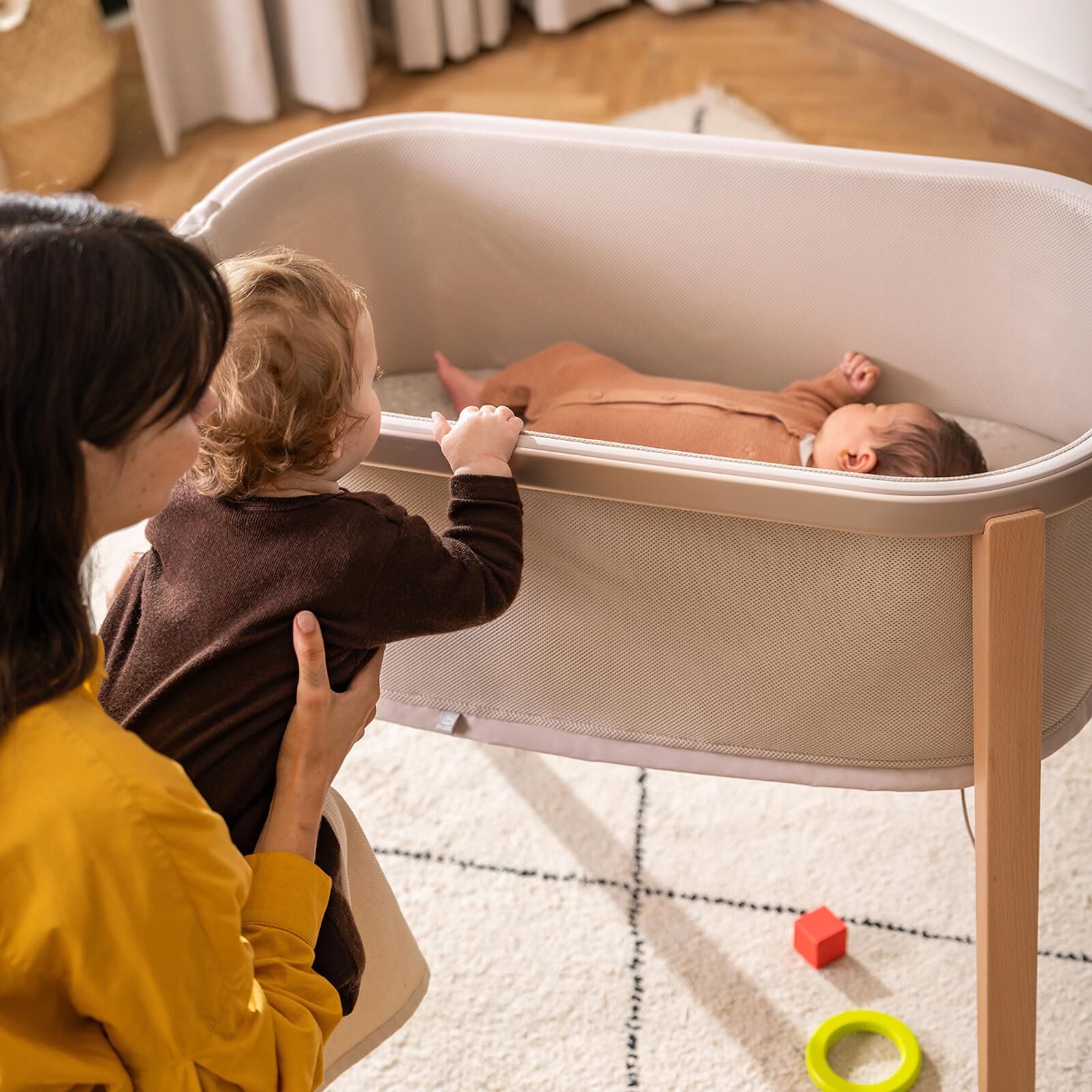 Mother holds child looking at newborn sleeping in a Stokke Nomi Bassinet.