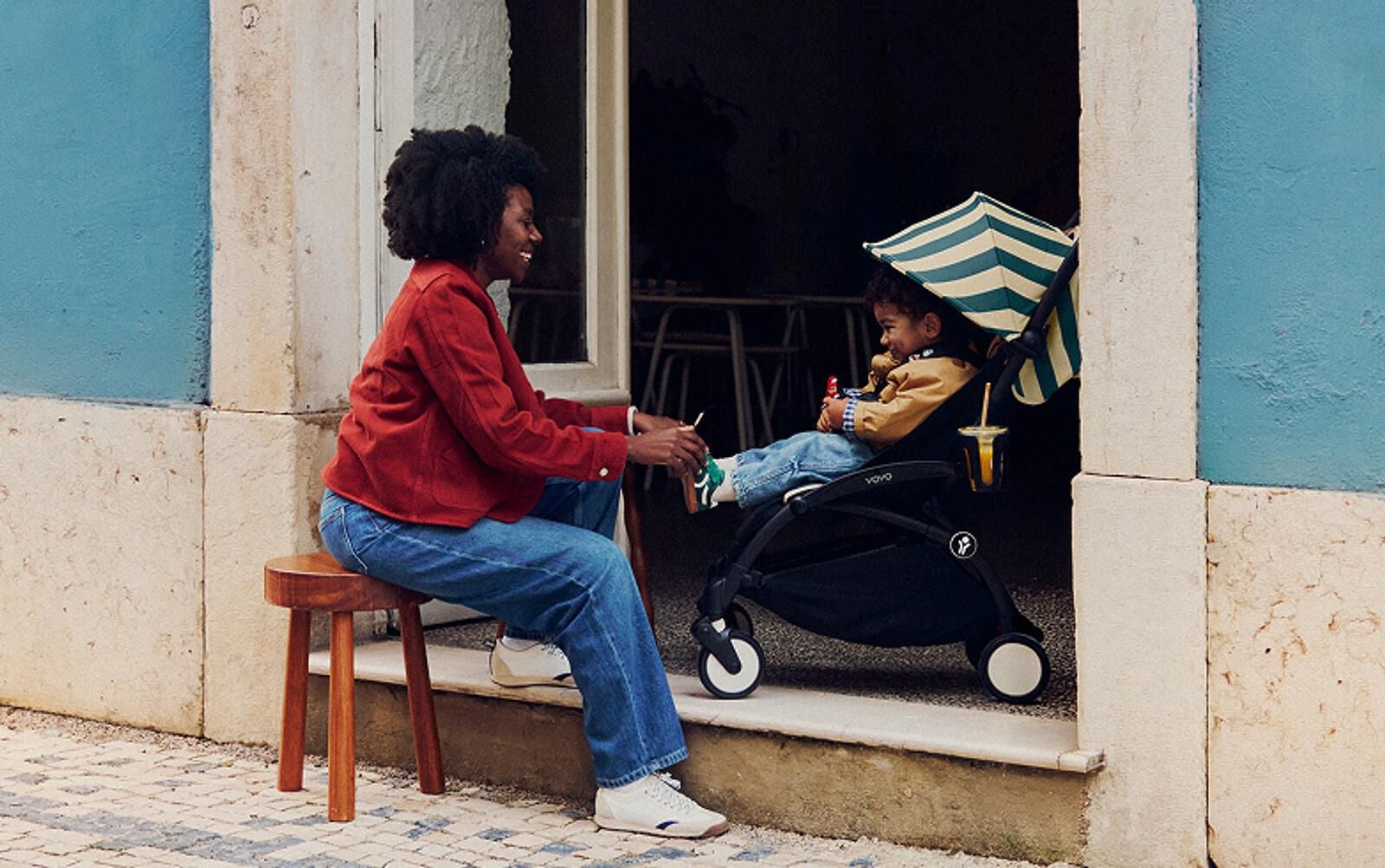 A smiling woman offers a drink to a child in a STOKKE YOYO stroller with a striped umbrella.