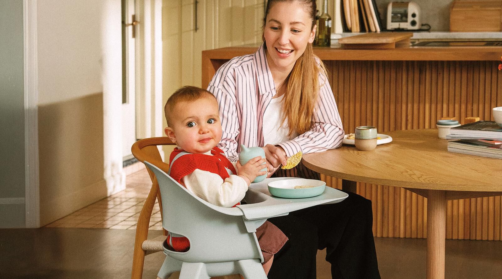 Happy baby in a high chair with a woman feeding them at a wooden table.