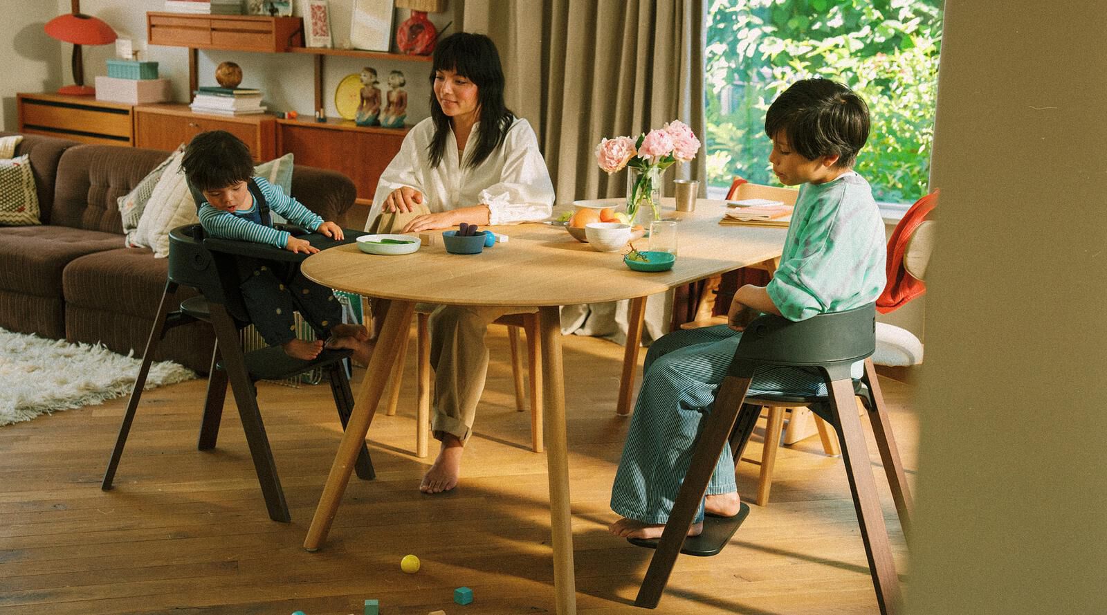 Een gezin van drie aan een lichte houten eettafel. Een kind in een donkere kinderstoel, een ander in een stoel.