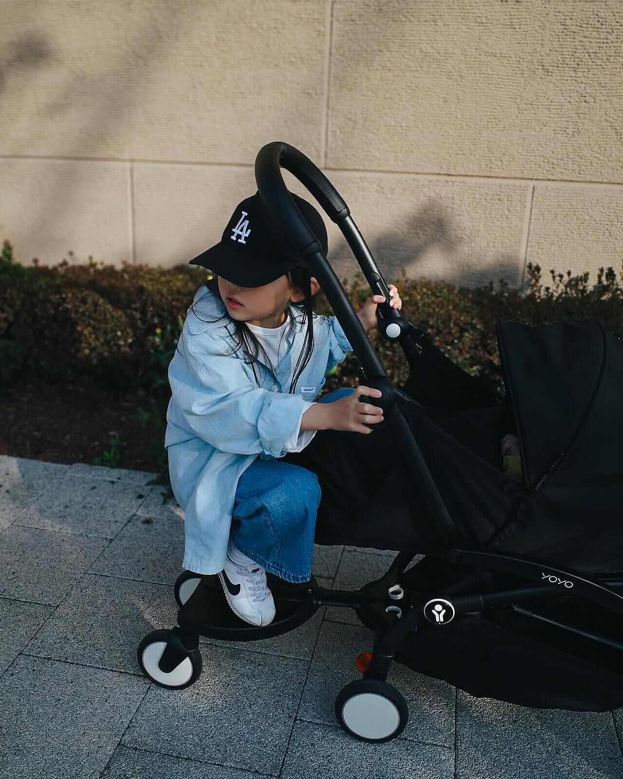 Child in LA cap stands on a wheeled board attached to a YOYO stroller.